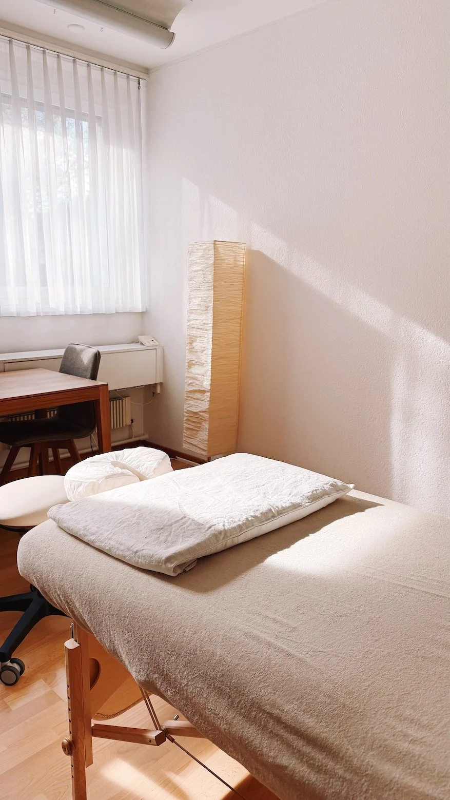 Picture of a massage bed in a sunlit room with a table, a chair and a lamp in the background