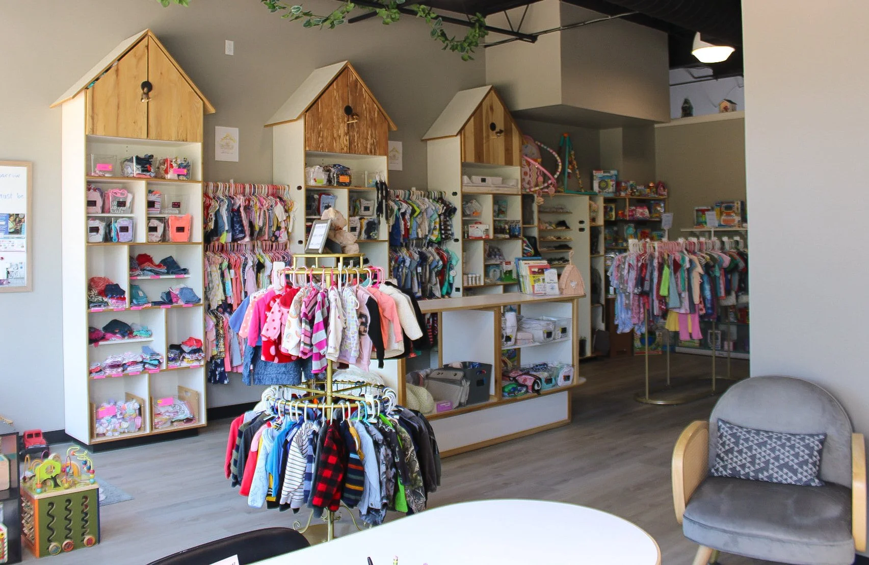 Interior of a children's clothing consignment store displaying racks of colorful children's clothing, shelves with toys and accessories, a cozy gray armchair with a patterned pillow, and a small round table.