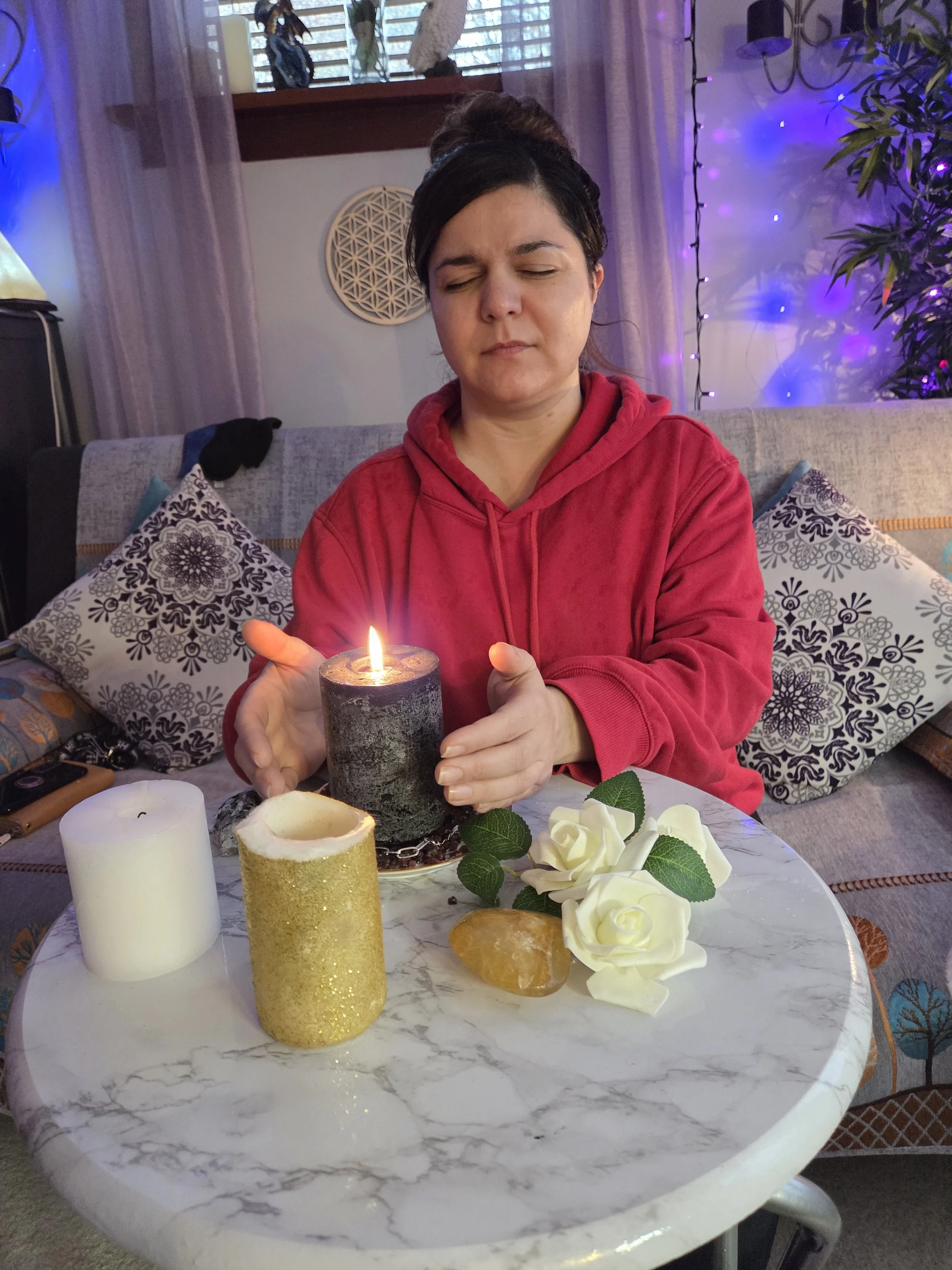 A woman in a red hoodie sitting at a marble table with burning candles, artificial white roses, a yellow crystal, and a white candle. She appears to be concentrating on the black candle in front of her, with closed eyes. The background includes a decorated living room with purple and blue lighting, patterned pillows, plants, and fairy lights.
