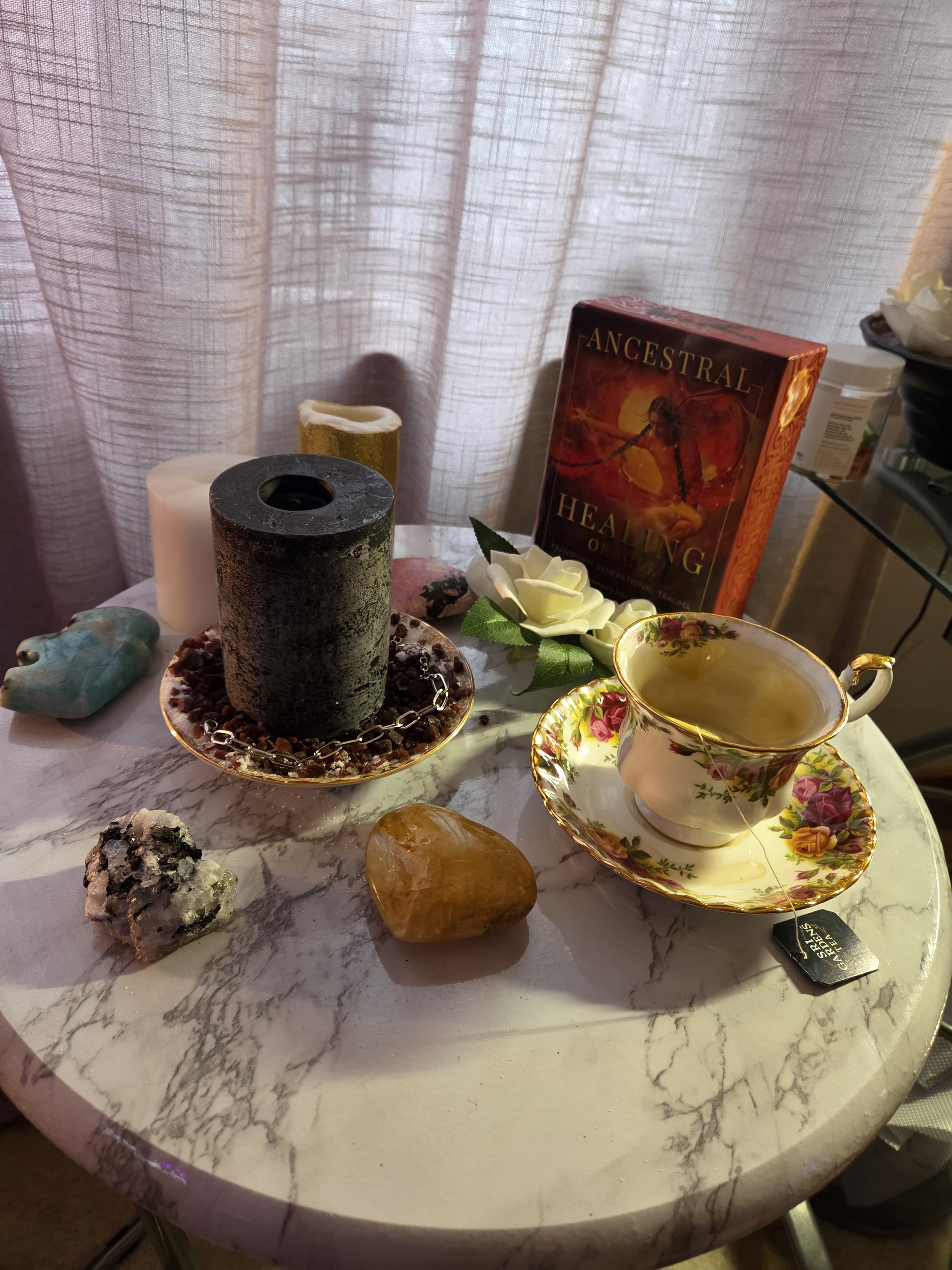 A round marble table holding a tea cup with floral and gold accents, a box of Ancestral Healing Oracle cards, a white rose, two candles (one black and one white), and various crystals, with sheer curtains in the background.