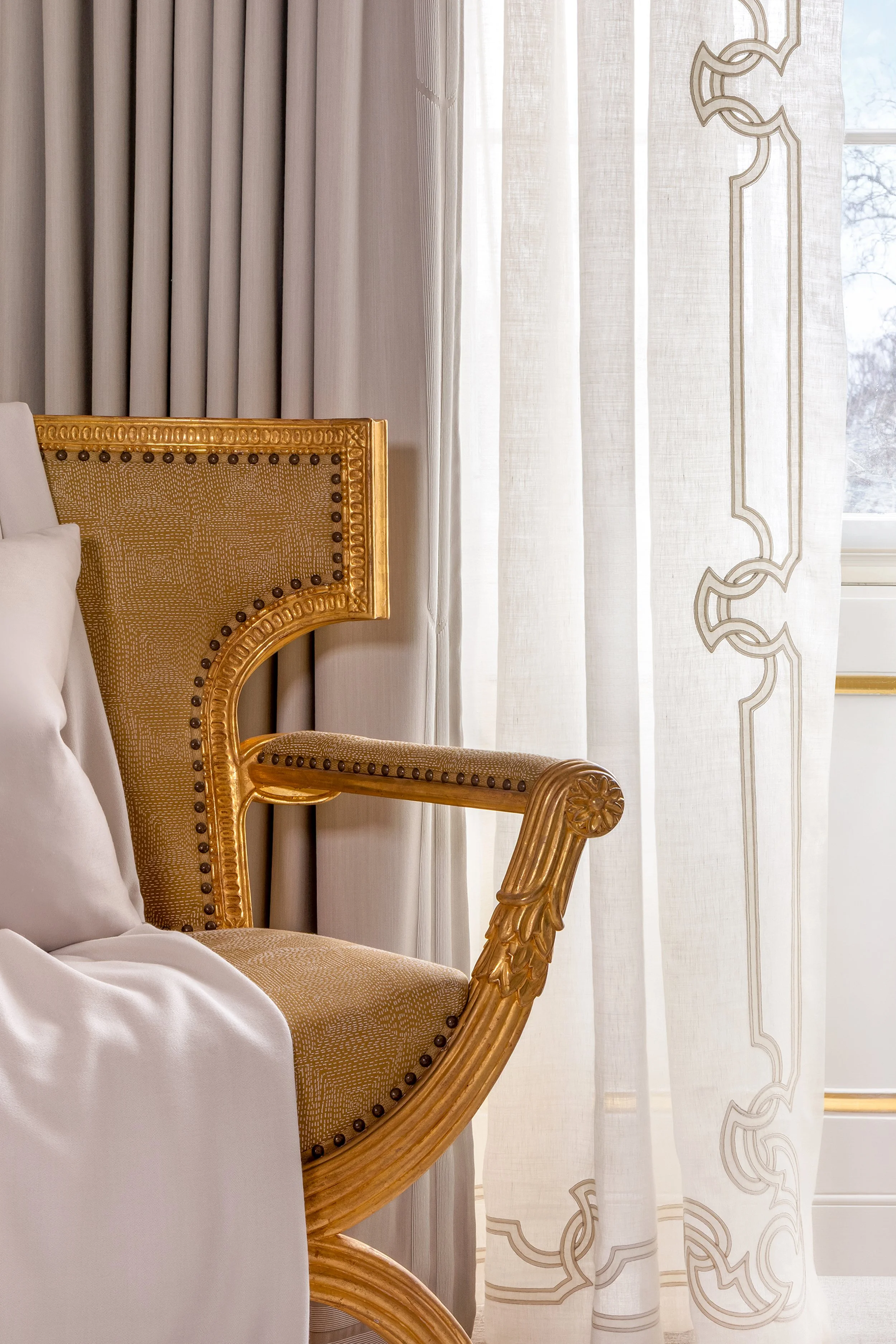 Close-up of an elegant gold-armrest chair next to white curtains and a window, with a pillow on the seat.