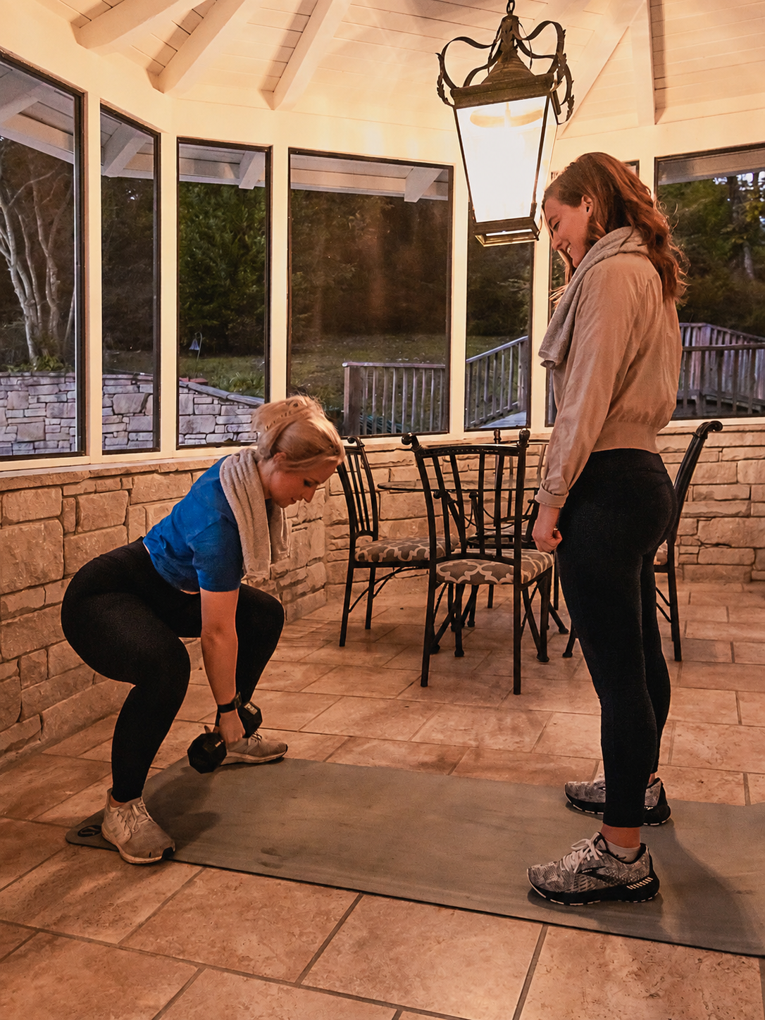 Two women during a HOMEFIT personal training session wearing cooling towels around their necks while strength training with dumbbells on a covered patio.