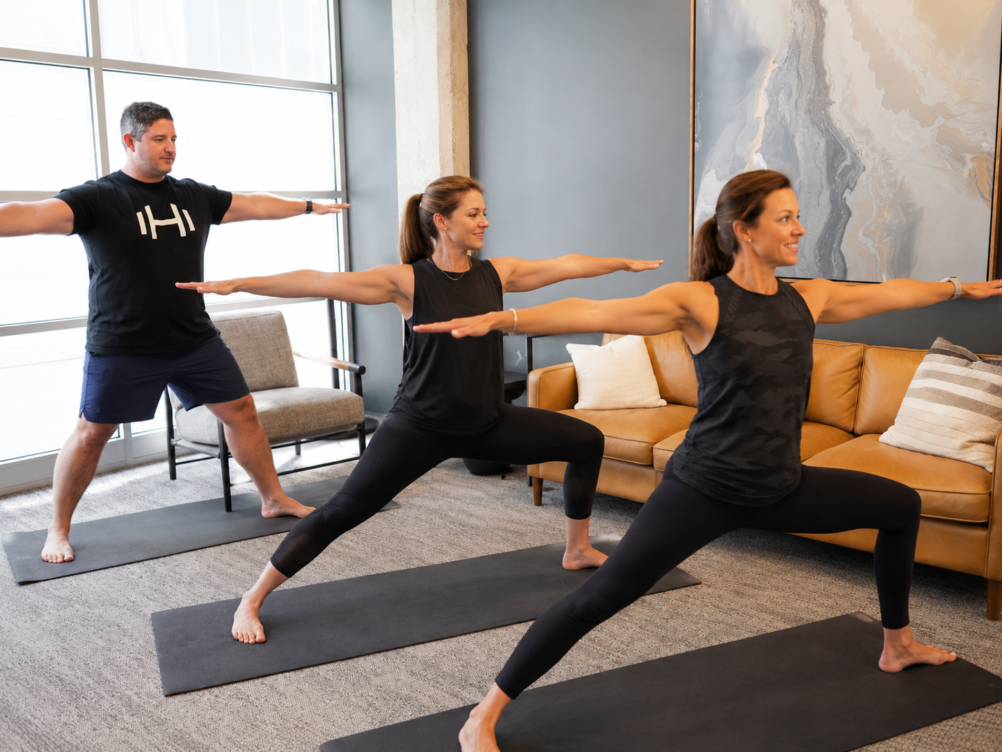 Three adults performing Warrior II yoga poses on yoga mats in a bright, modern living room. Large windows, contemporary furnishings, and an abstract wall art piece create a welcoming space for a guided yoga session.