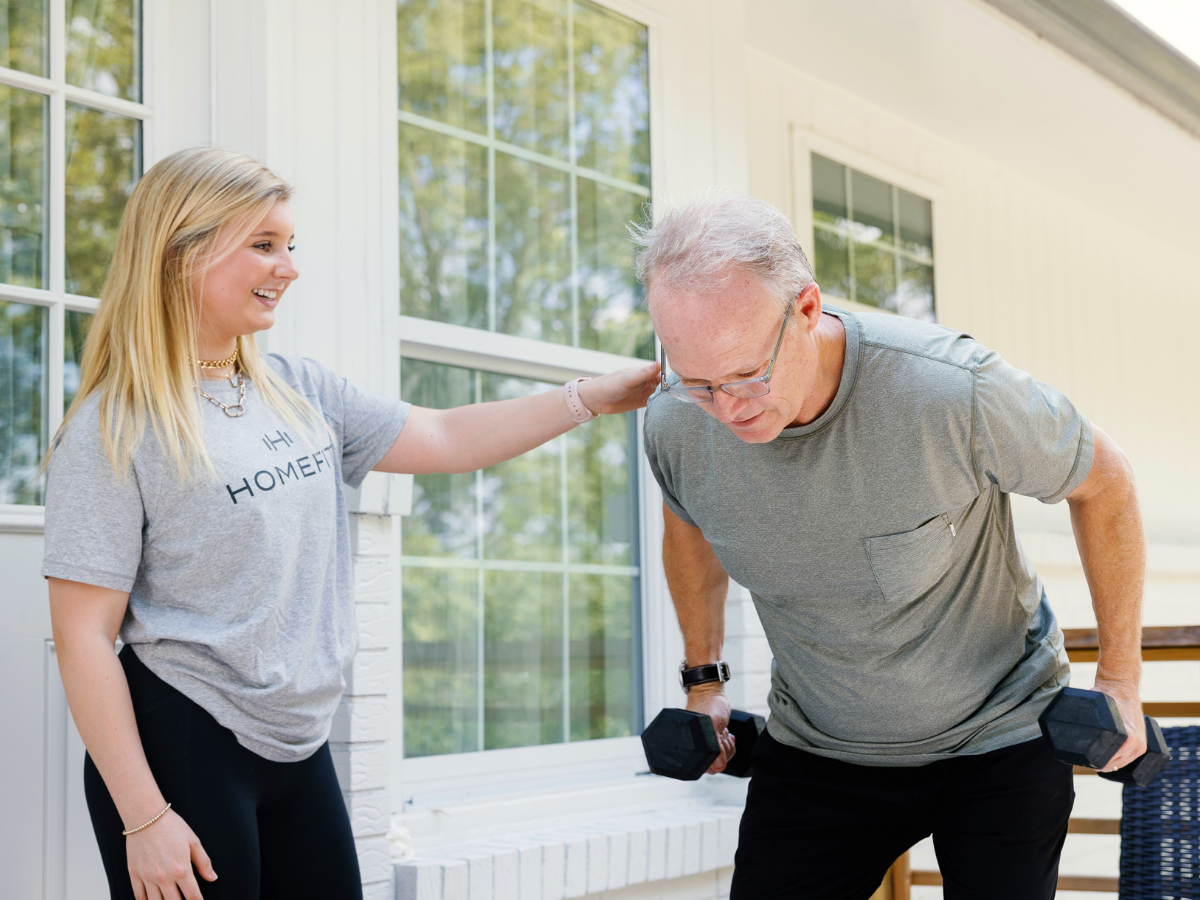 HOMEFIT trainer supporting an older adult client during an outdoor dumbbell workout at home, showcasing personalized in-home fitness training for strength and mobility.