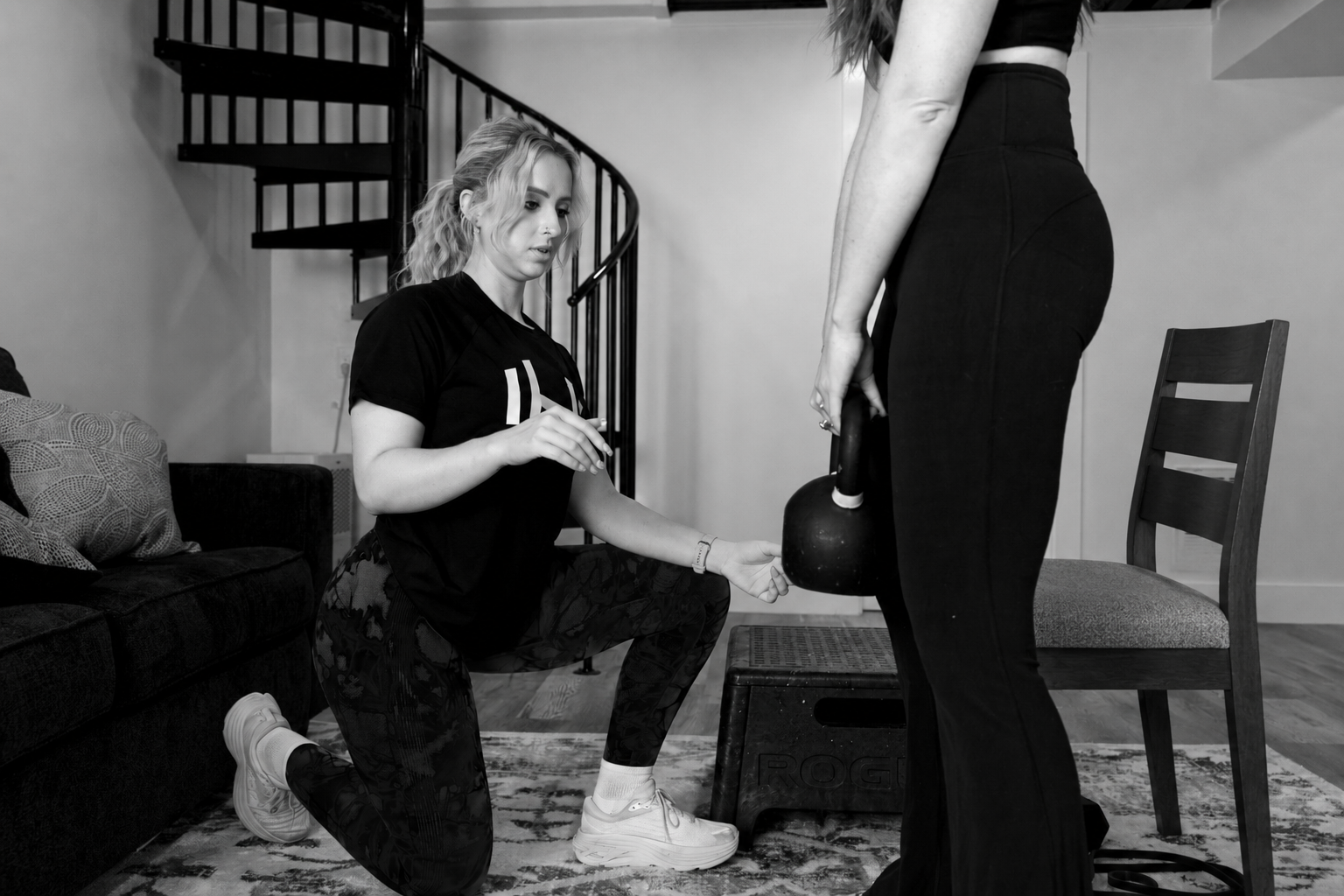 HOMEFIT personal trainer coaching a woman through a kettlebell exercise beside a sturdy chair during an in-home workout.