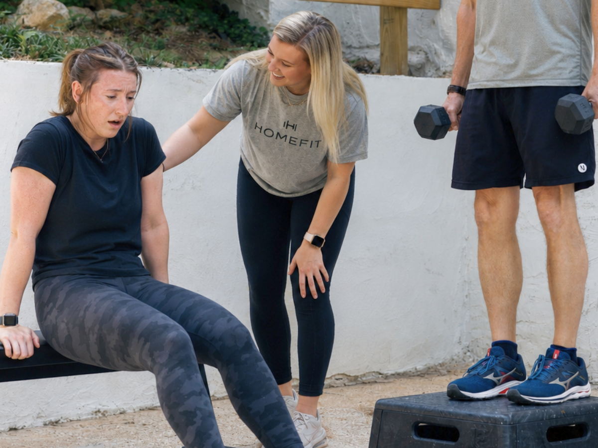 HOMEFIT trainer supports a tired client during an outdoor workout while another client stands nearby holding dumbbells on a step platform.