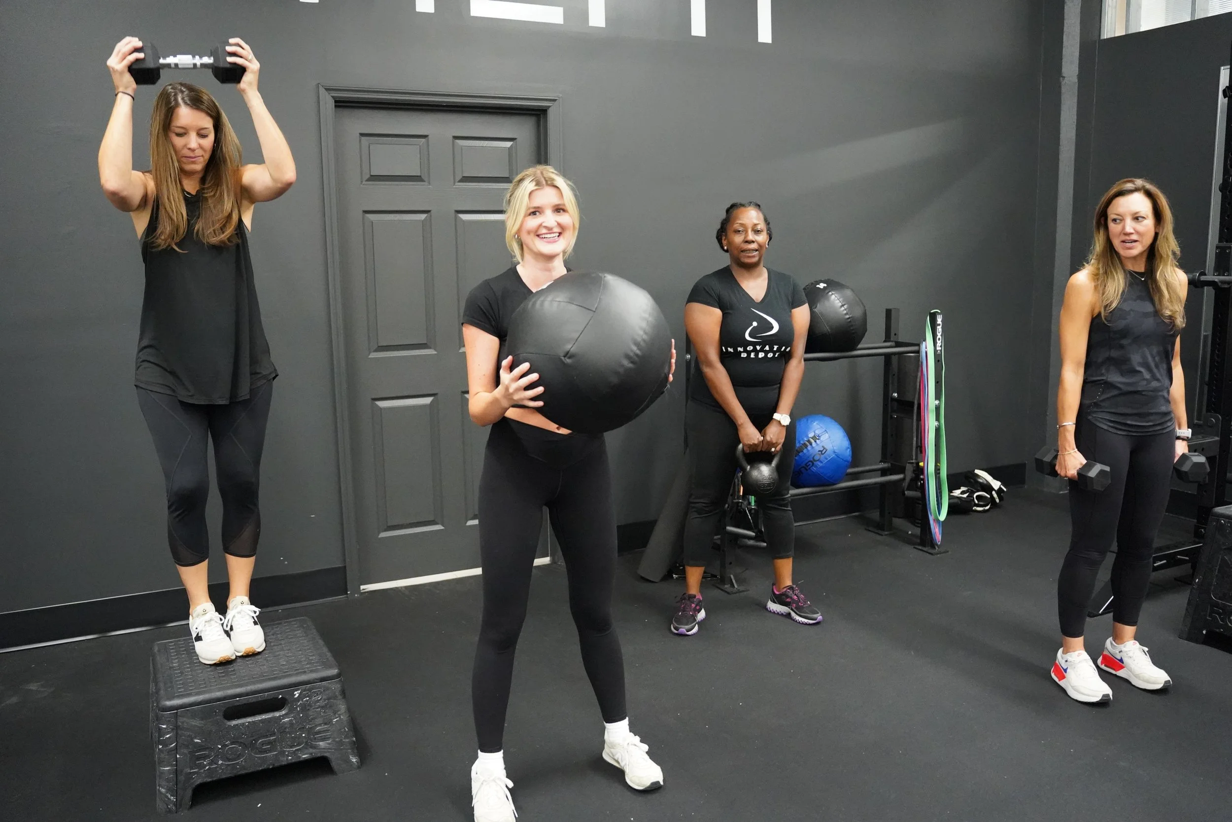 Group fitness training session featuring women performing strength exercises with dumbbells, medicine balls, and kettlebells, highlighting HOMEFIT's personalized small-group workouts and social fitness benefits.