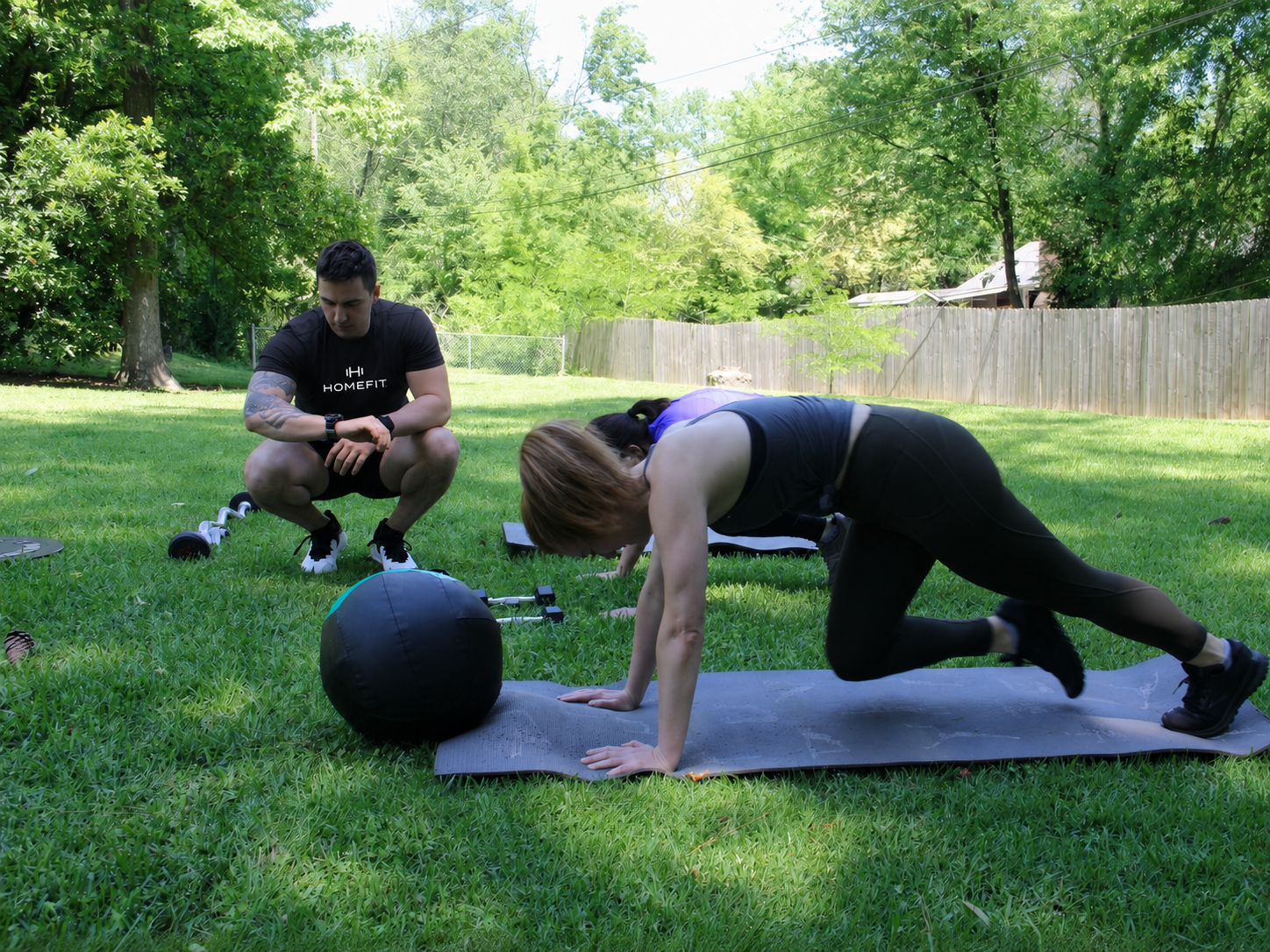 HOMEFIT trainer monitors an outdoor backyard workout as two women perform mat exercises with fitness equipment nearby.