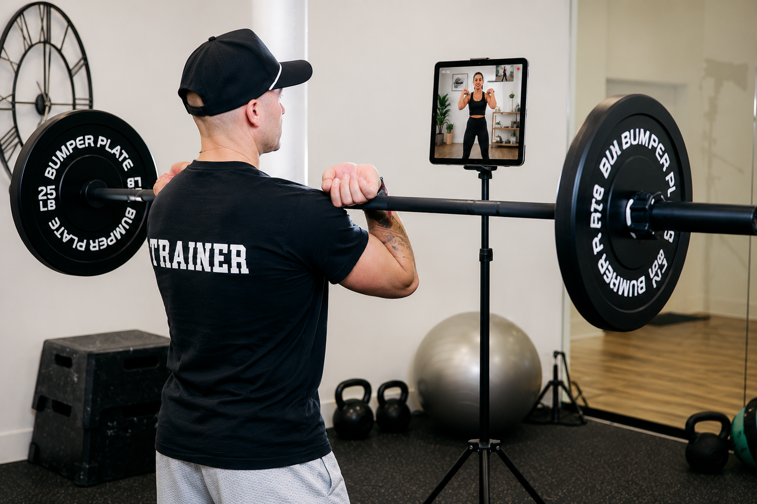 Personal trainer performing a barbell front-rack exercise while following a live virtual training session on a tablet, demonstrating online fitness coaching and remote workout support.