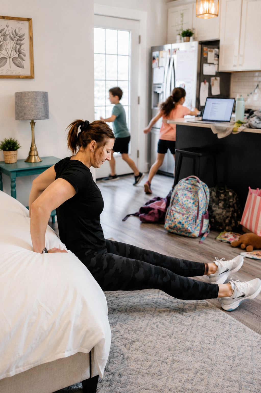 Woman doing triceps dips at home beside a bed while kids move through the kitchen in the background, showing a busy real-life home workout moment.