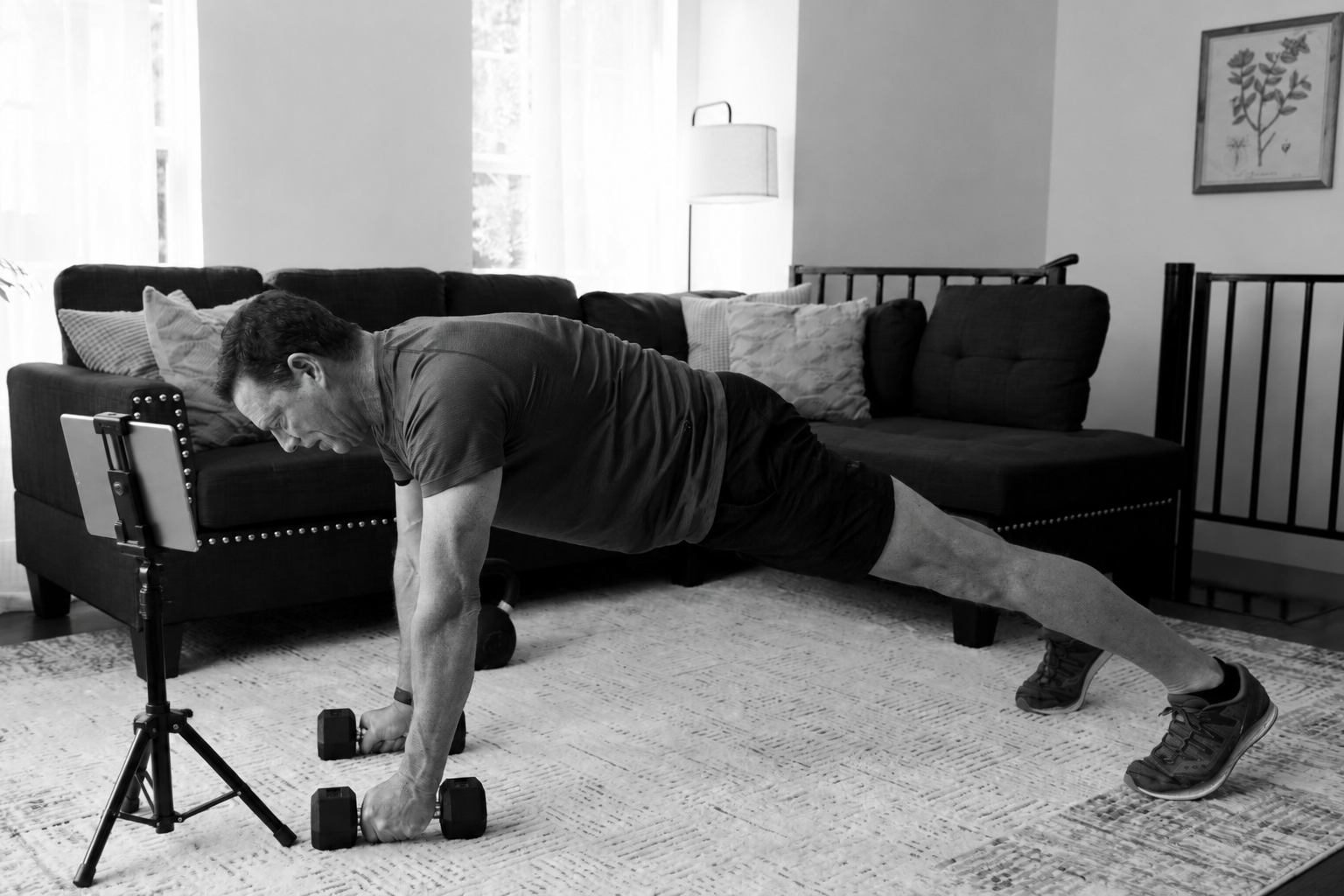 Man performing a plank exercise with dumbbells in a bright living room while following a virtual personal training session on an iPad mounted to a tripod.