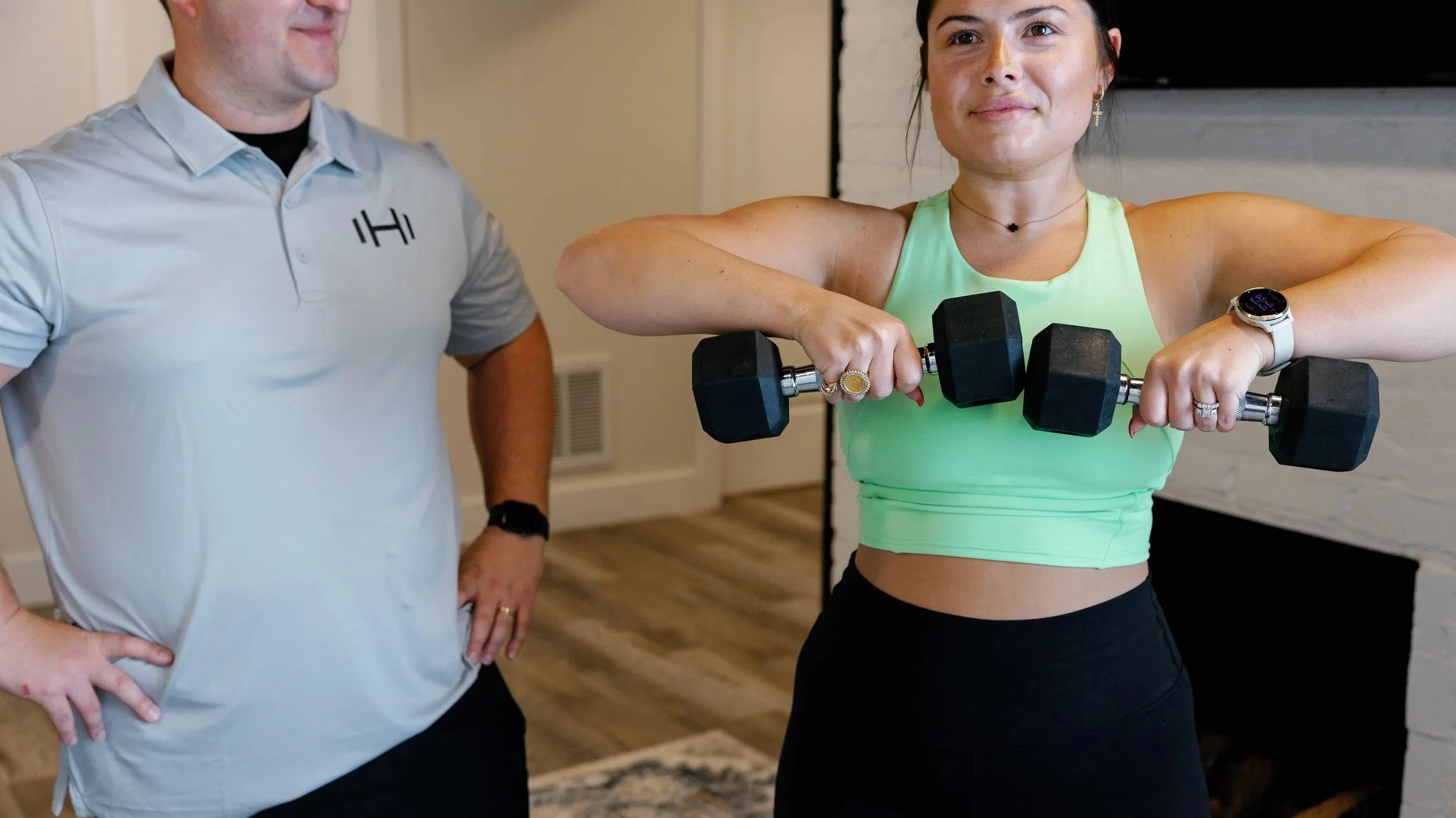 A woman performs a controlled upright row with dumbbells while a trainer observes and guides nearby.