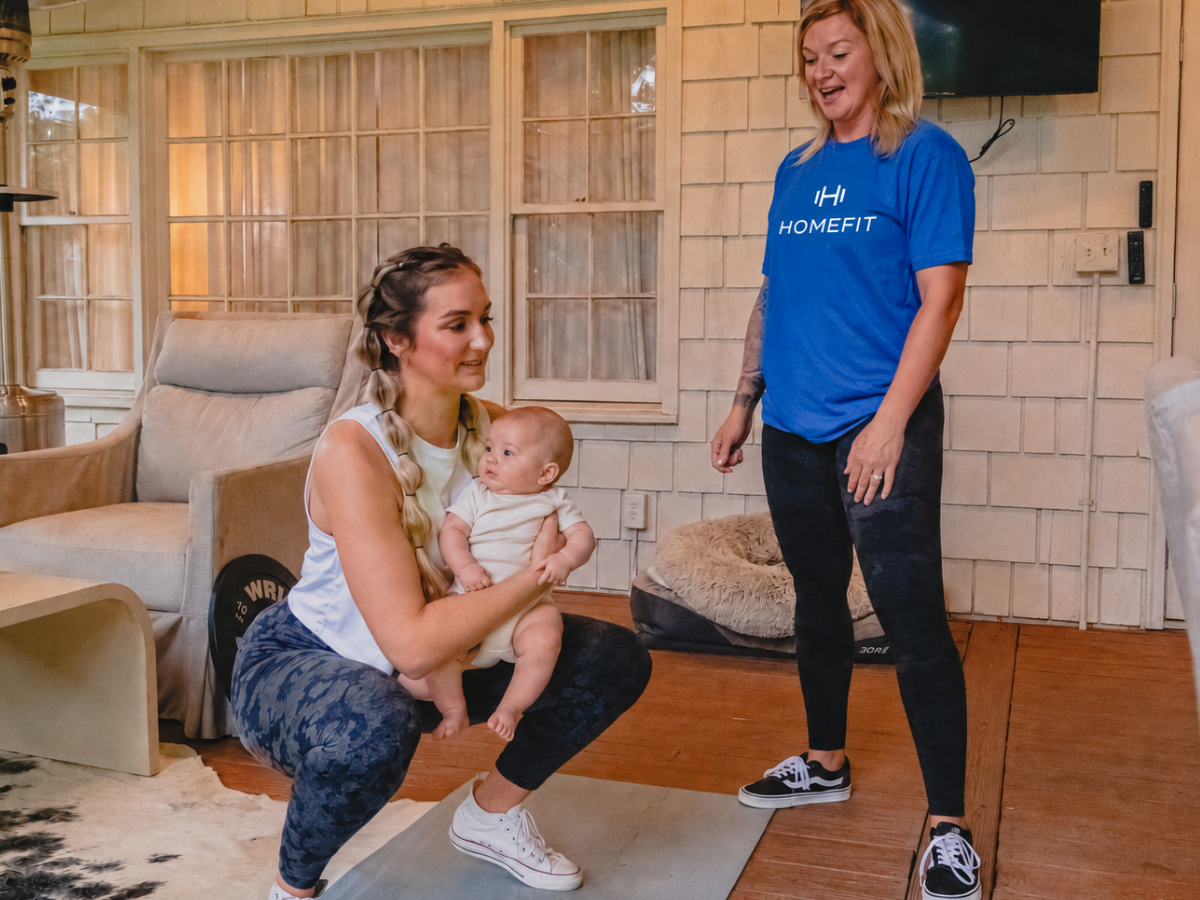 New mom performing a supported squat while holding her baby as a HOMEFIT postpartum personal trainer provides guidance at home.
