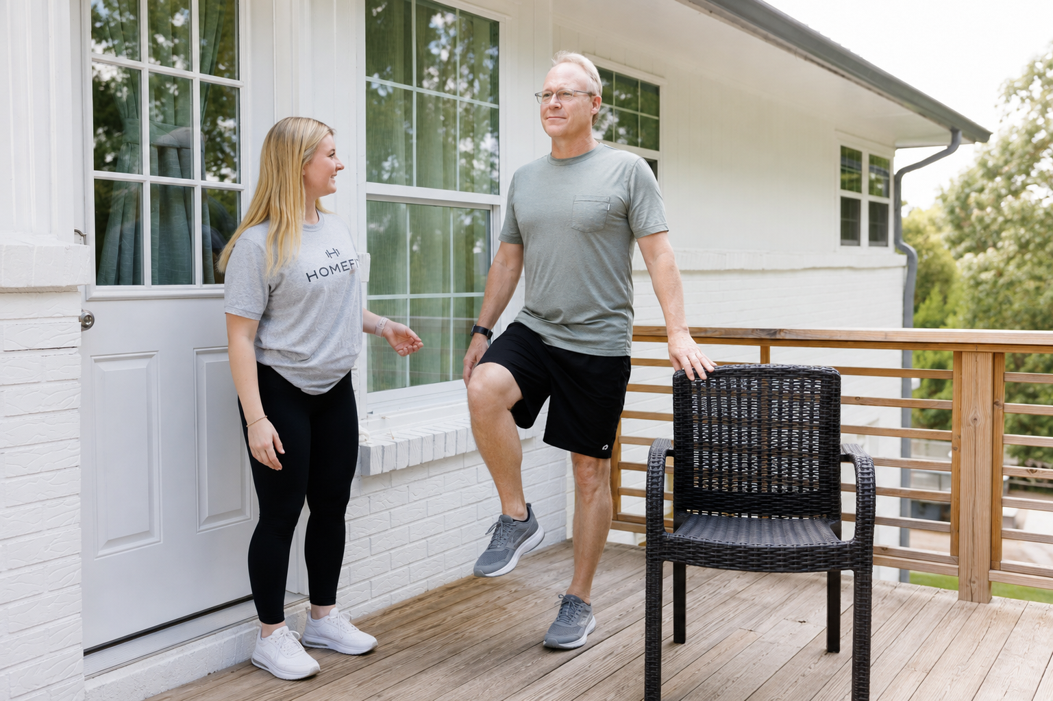 Older adult practicing a controlled single-leg balance exercise with a HOMEFIT personal trainer and sturdy chair nearby for support.