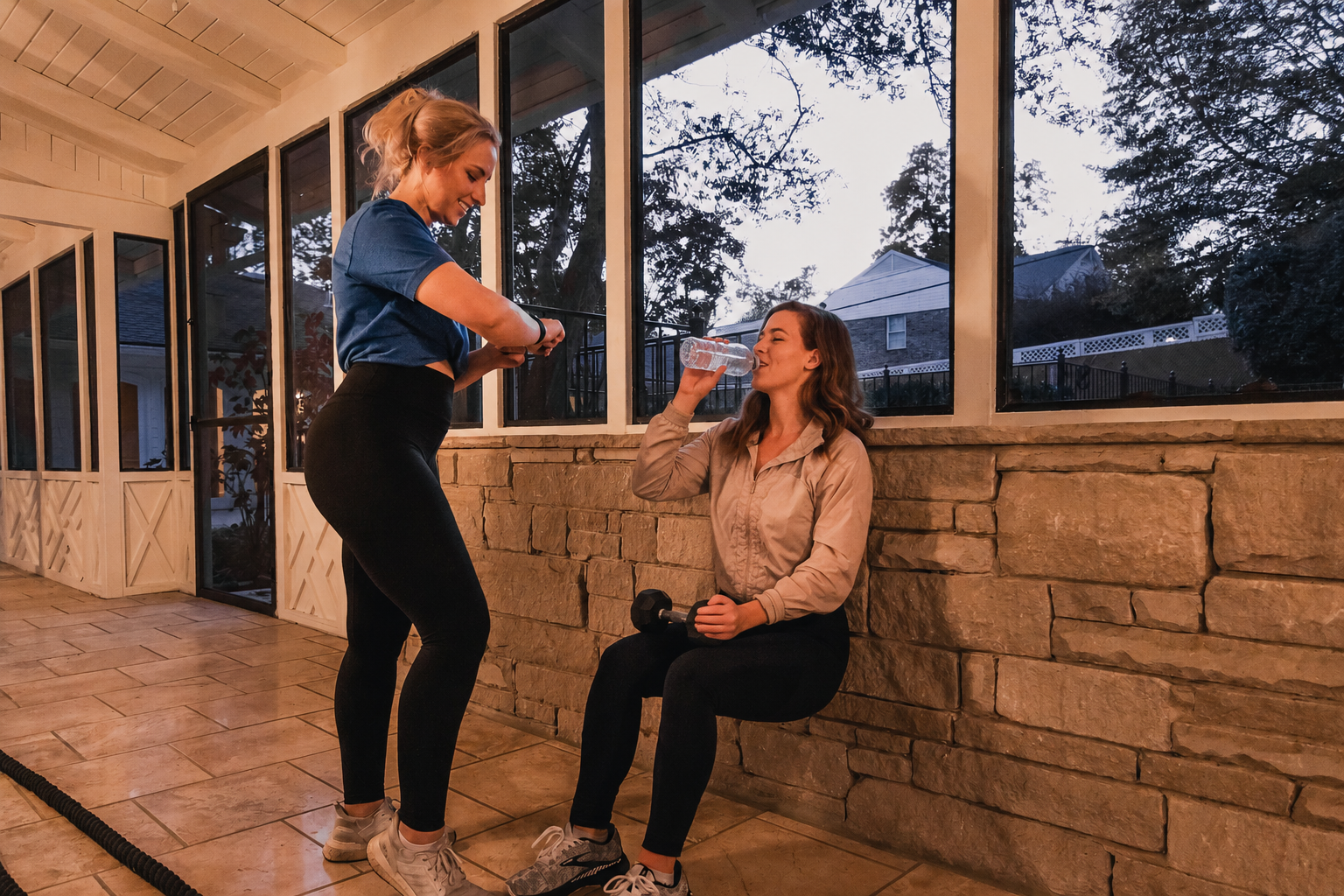 HOMEFIT client taking a water break after a workout while a trainer checks progress in a sunroom, highlighting hydration and recovery during personal training.