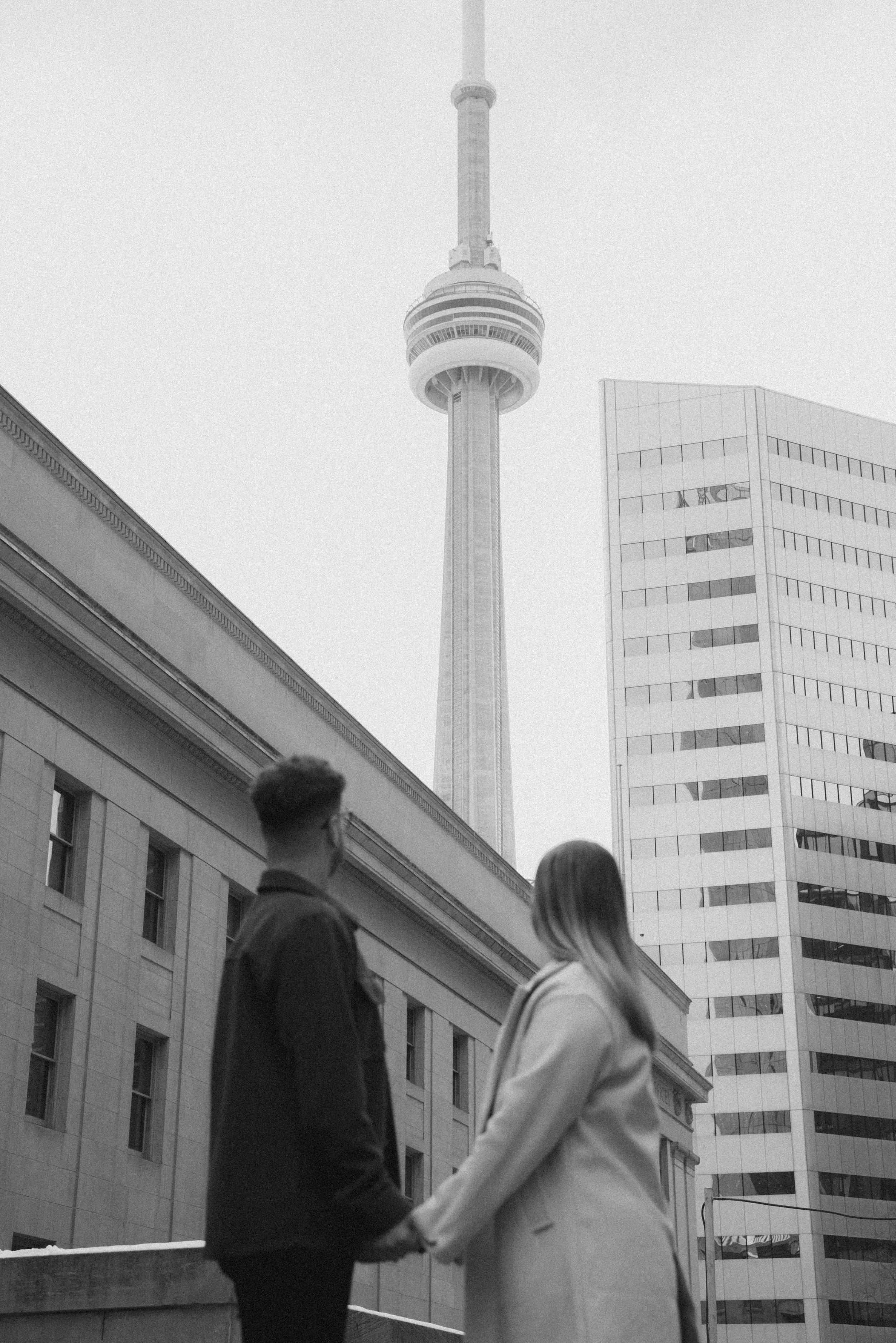 A couple holding hands on a city rooftop with the CN Tower and tall modern buildings in the background.