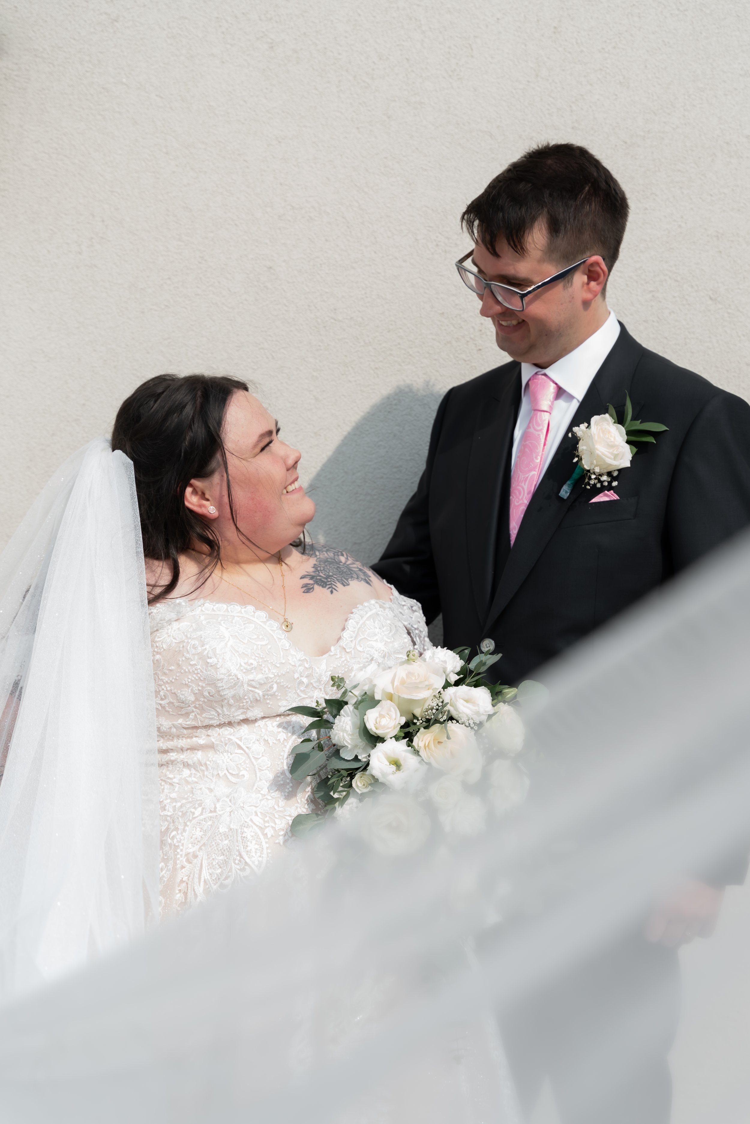 Bride and groom smiling at each other, with the bride holding a bouquet of white roses, standing against a light-colored wall.