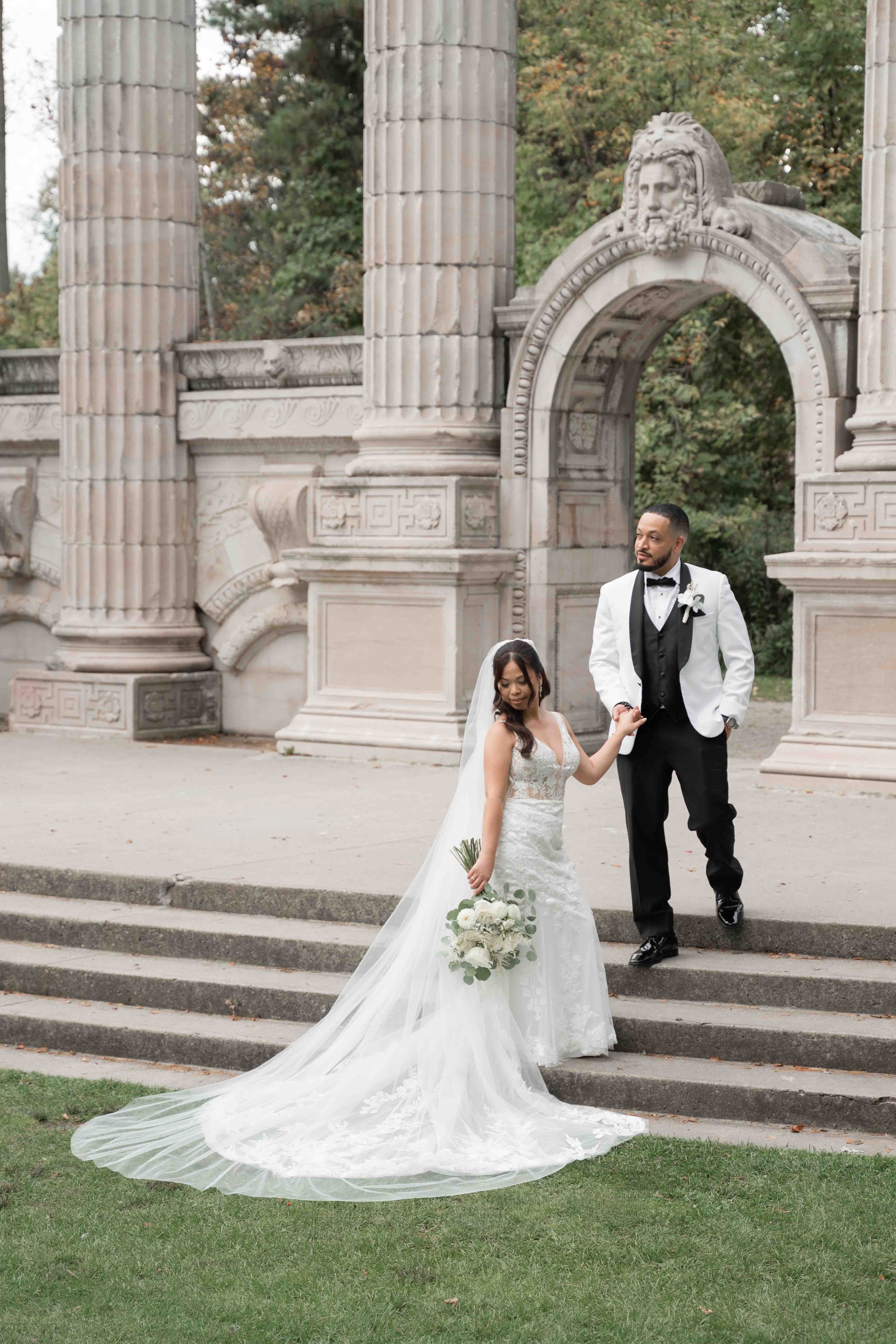 A bride and groom pose on steps in front of a historic stone archway, with the bride holding a bouquet of white roses and greenery, and the groom dressed in a white tuxedo jacket with a black vest and bow tie.