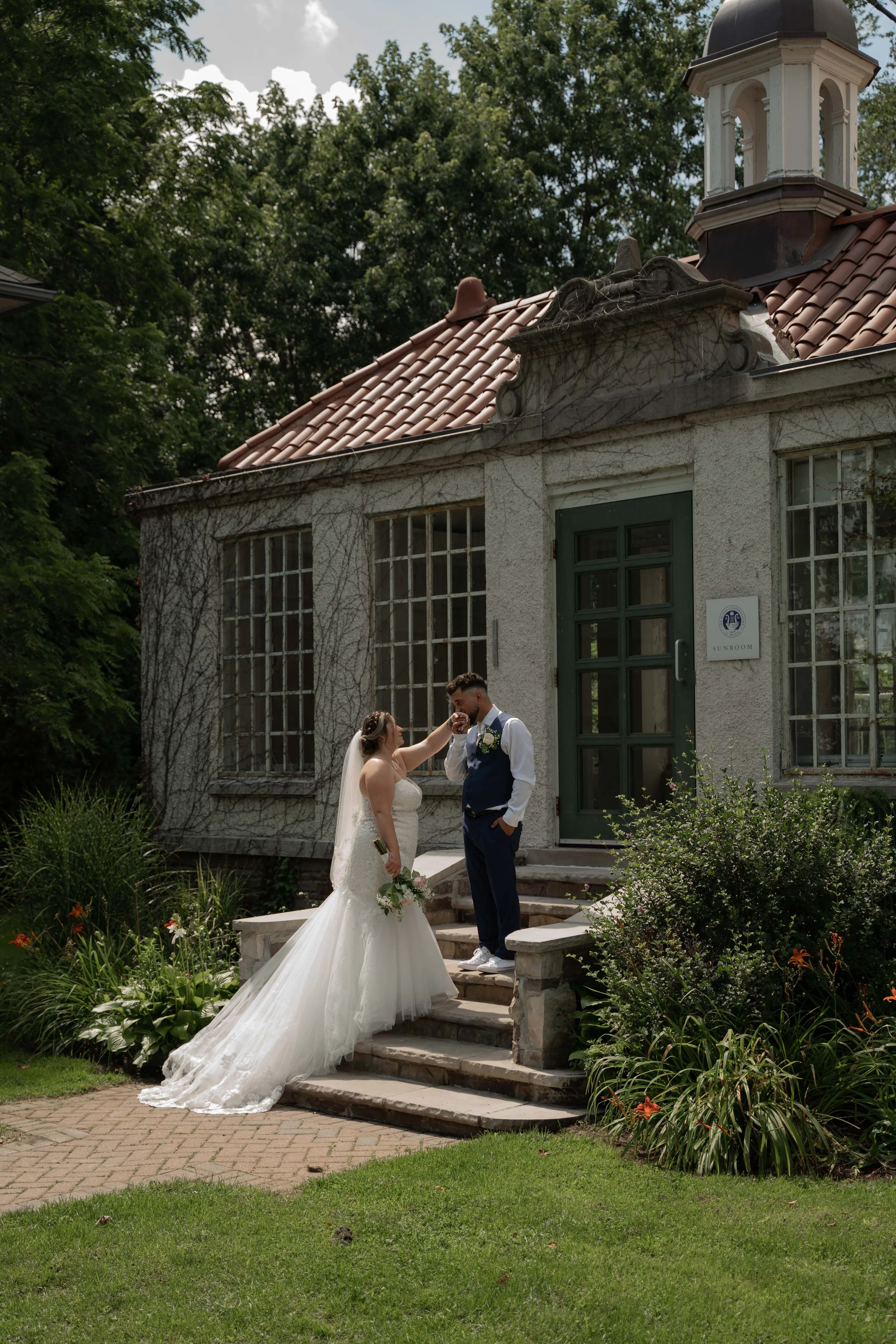 Bride and groom sharing a moment on the steps of a small, vine-covered building with a sign that says 'Sunroom.' The bride is in a white wedding dress, and the groom is in a dark suit with a white shirt. They are outside, surrounded by greenery and flowers.