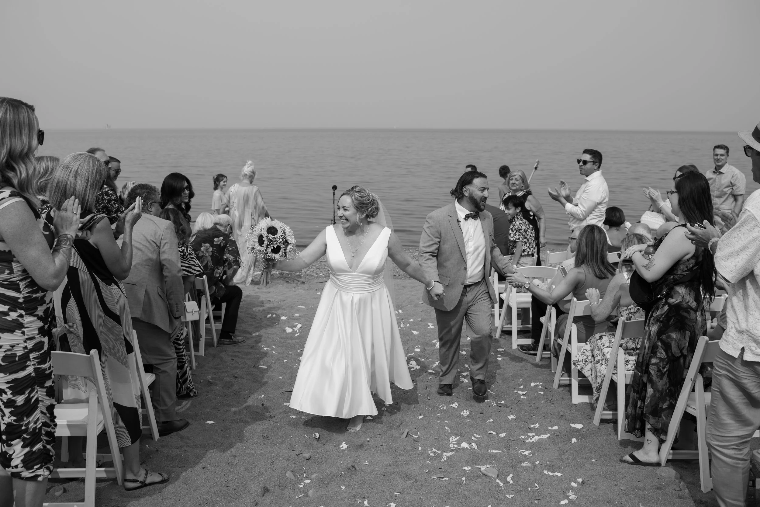 A wedding couple is walking hand in hand on a beach, smiling, with guests clapping and taking photos, during a wedding ceremony by the water.