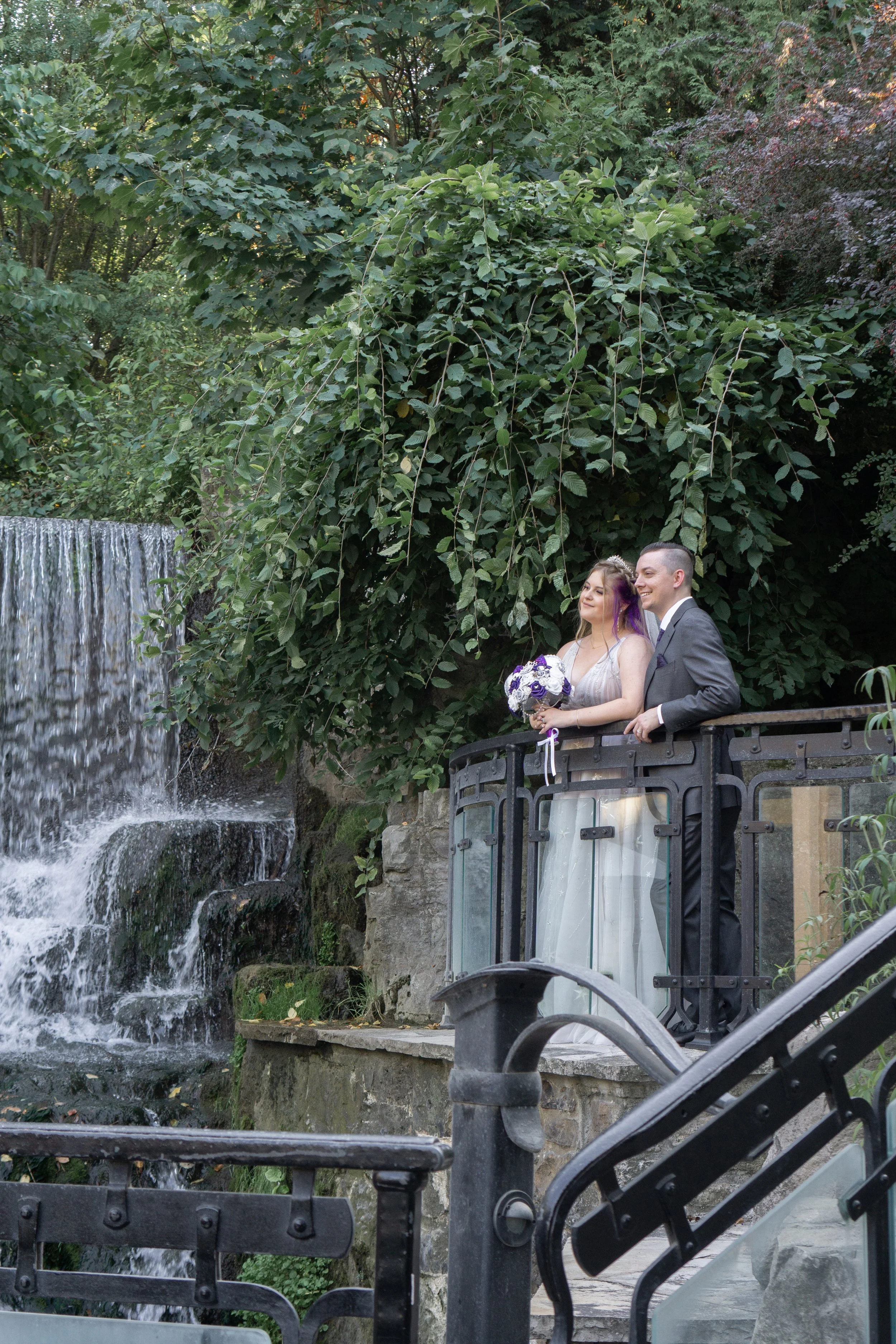 A newlywed couple stands on a small balcony overlooking a waterfall and lush greenery, with the bride holding a bouquet of purple and white flowers.