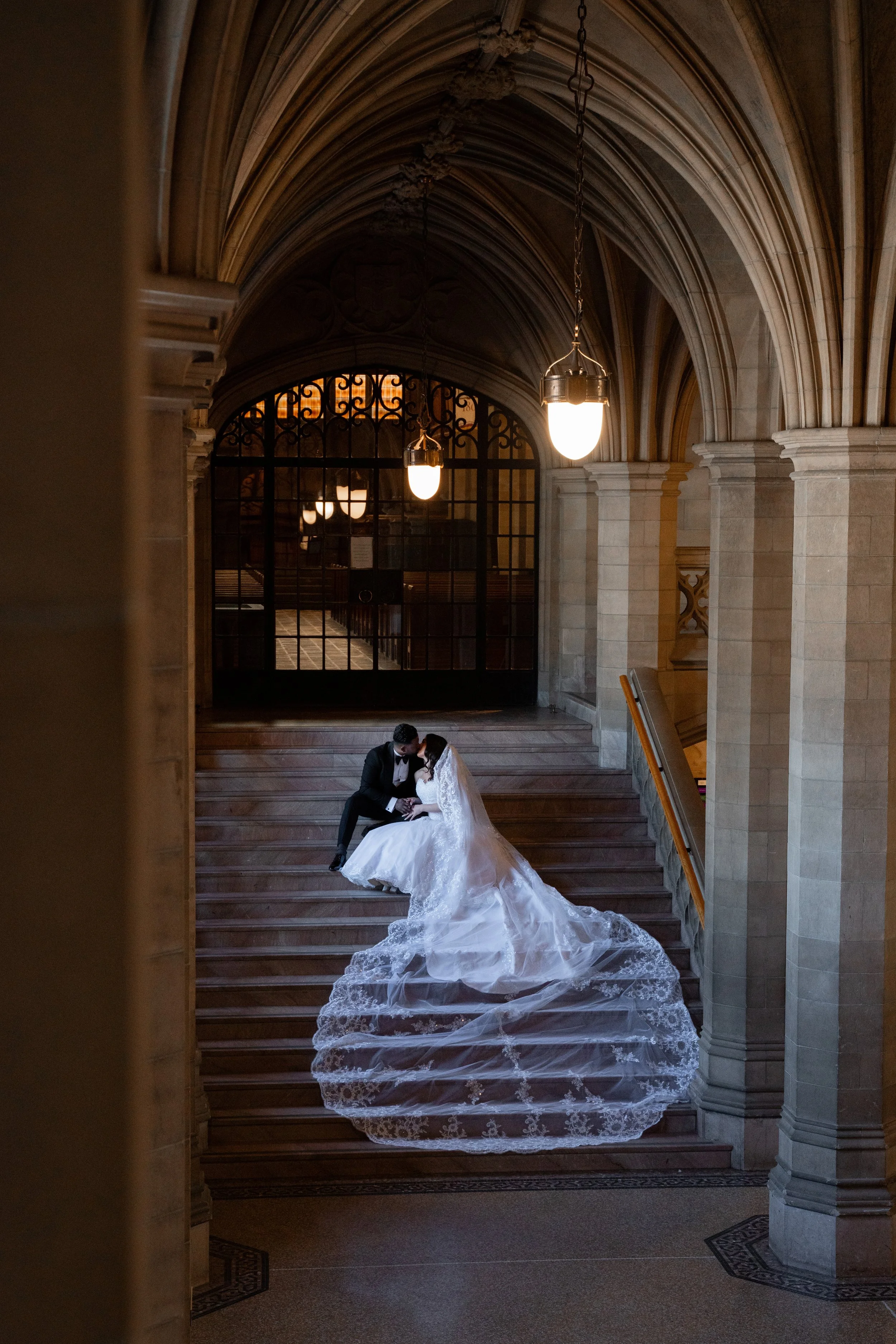 A bride and groom wearing wedding attire sitting on a grand staircase inside a historic building with arched ceilings and hanging lights, sharing an intimate moment.