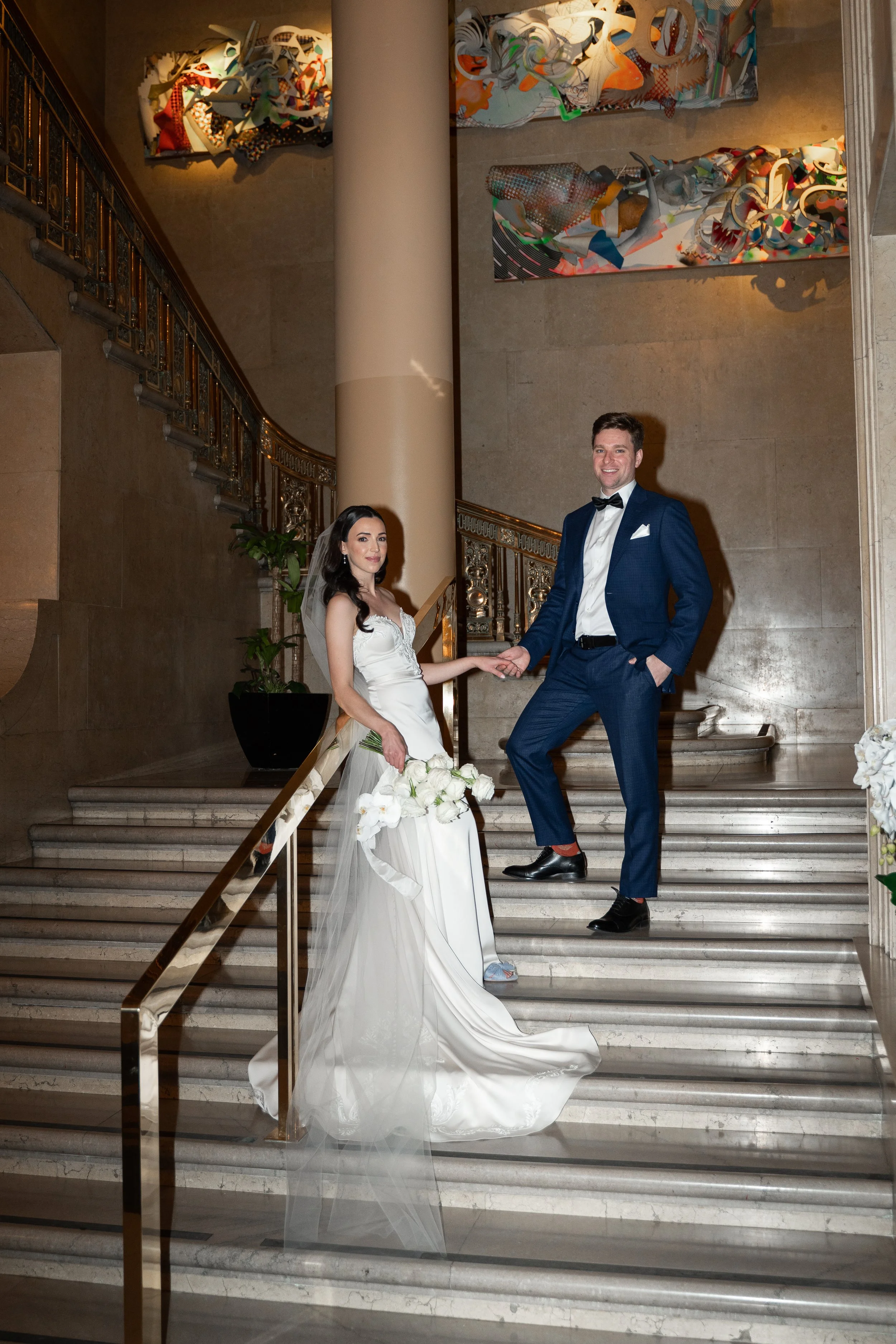 A bride and groom standing on a staircase inside a building, holding hands. The bride is wearing a white wedding gown and holding a bouquet of white flowers, while the groom is dressed in a navy blue suit with a bow tie.