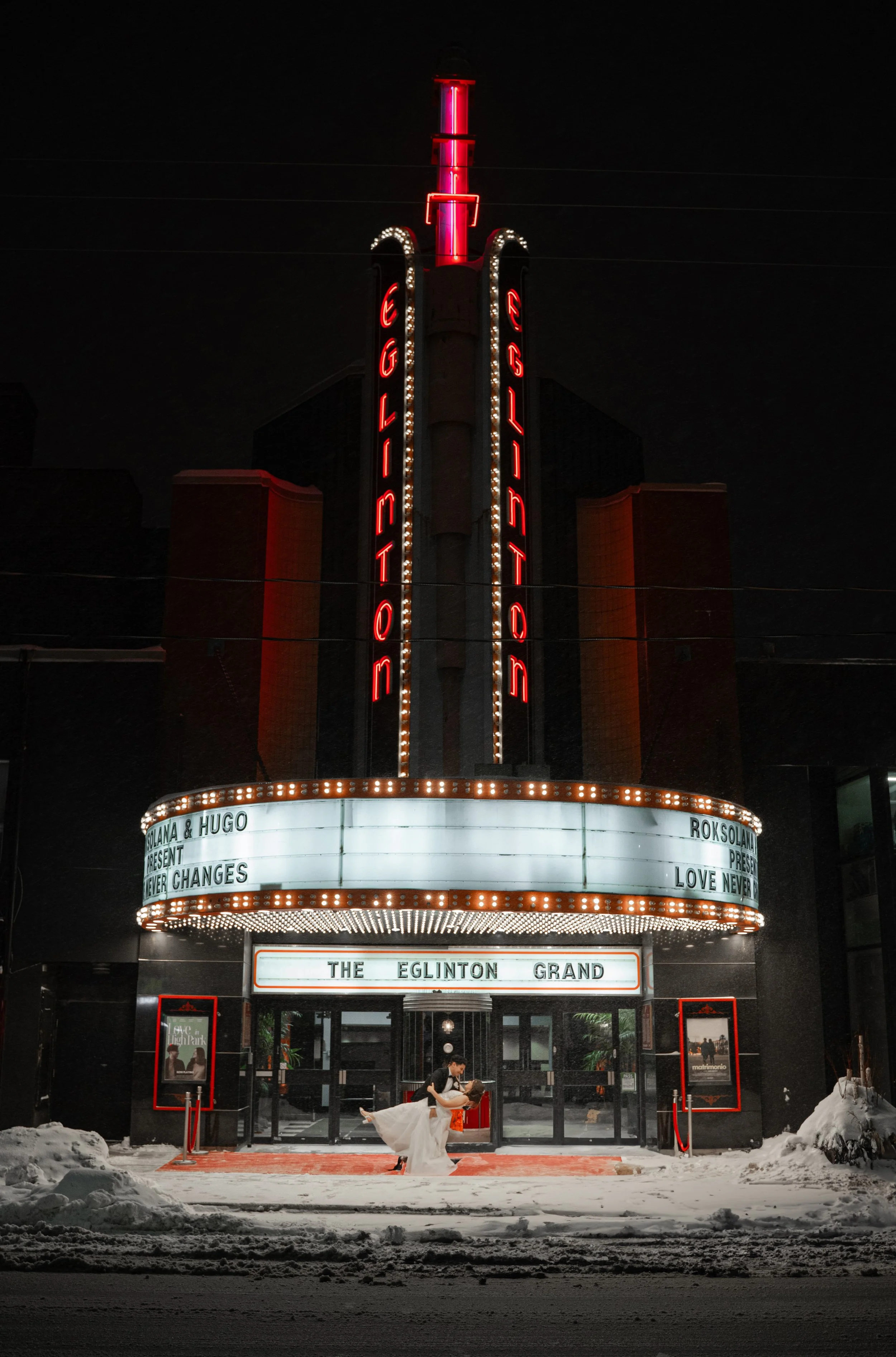 A couple dancing in front of a theater marquee at night, with snow on the ground, theater sign reading 'The Eglinton Grand', and neon lights and signage on the building.