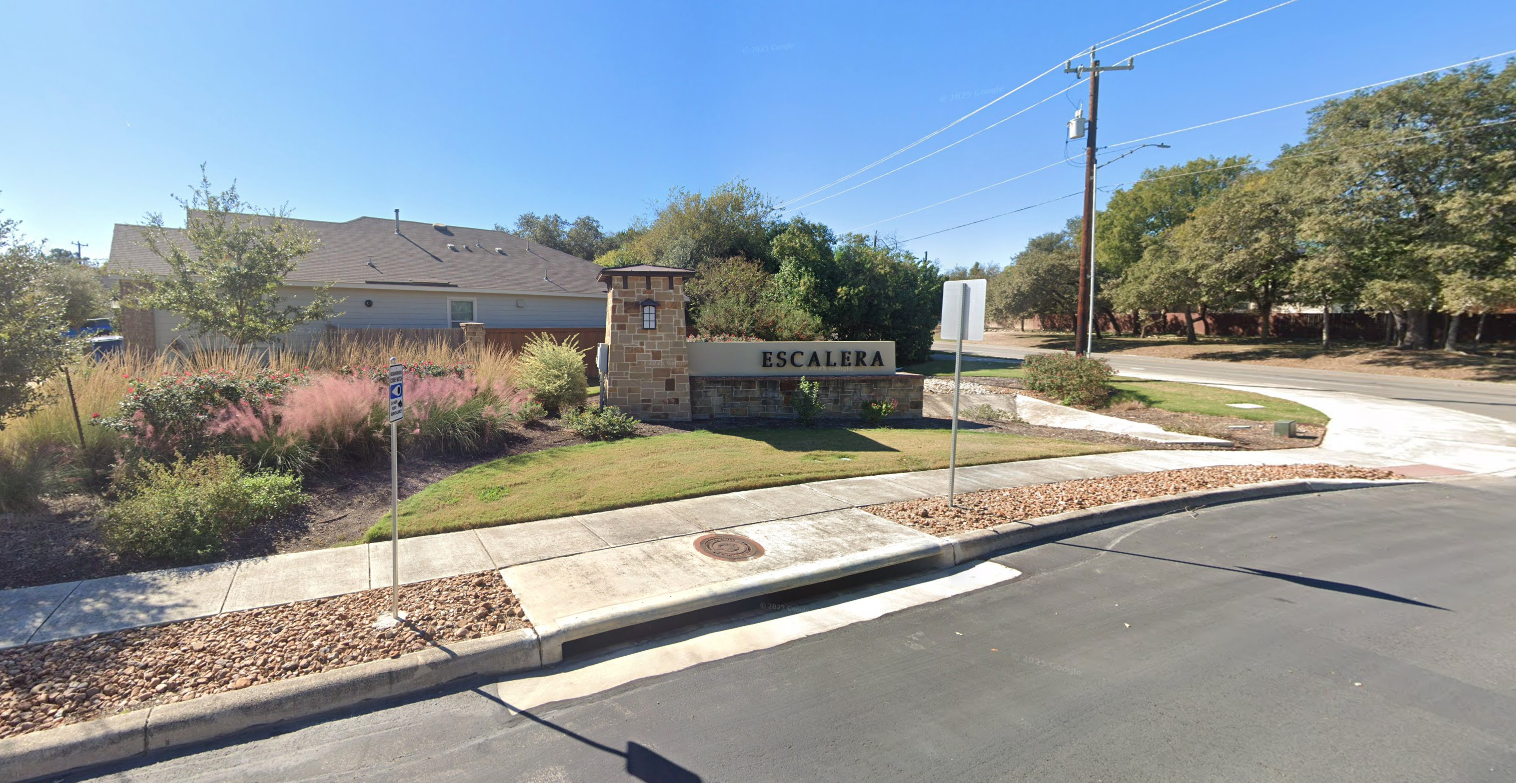 Street view of the entrance to Escalera neighborhood with a stone sign, landscaped bushes, and a sidewalk. There are trees, power lines, and a house in the background under a clear blue sky.