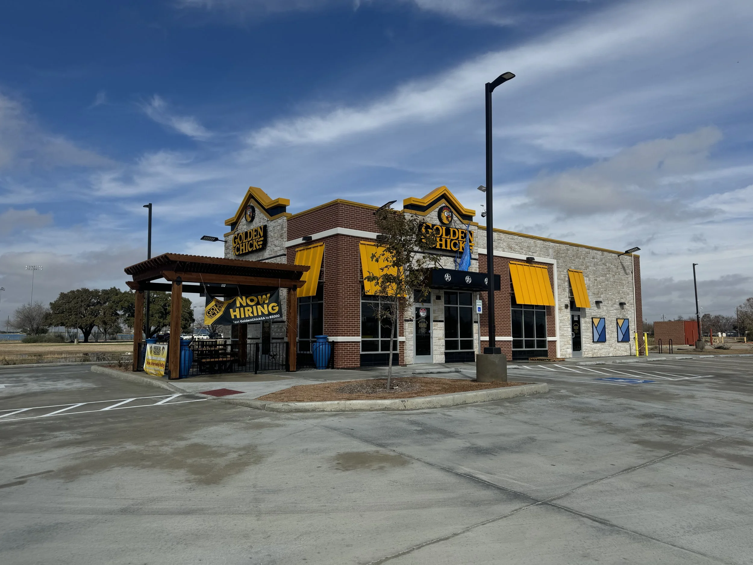 Empty Golden Chick restaurant with 'Now Hiring' sign, parked in a largely empty lot under partly cloudy sky.