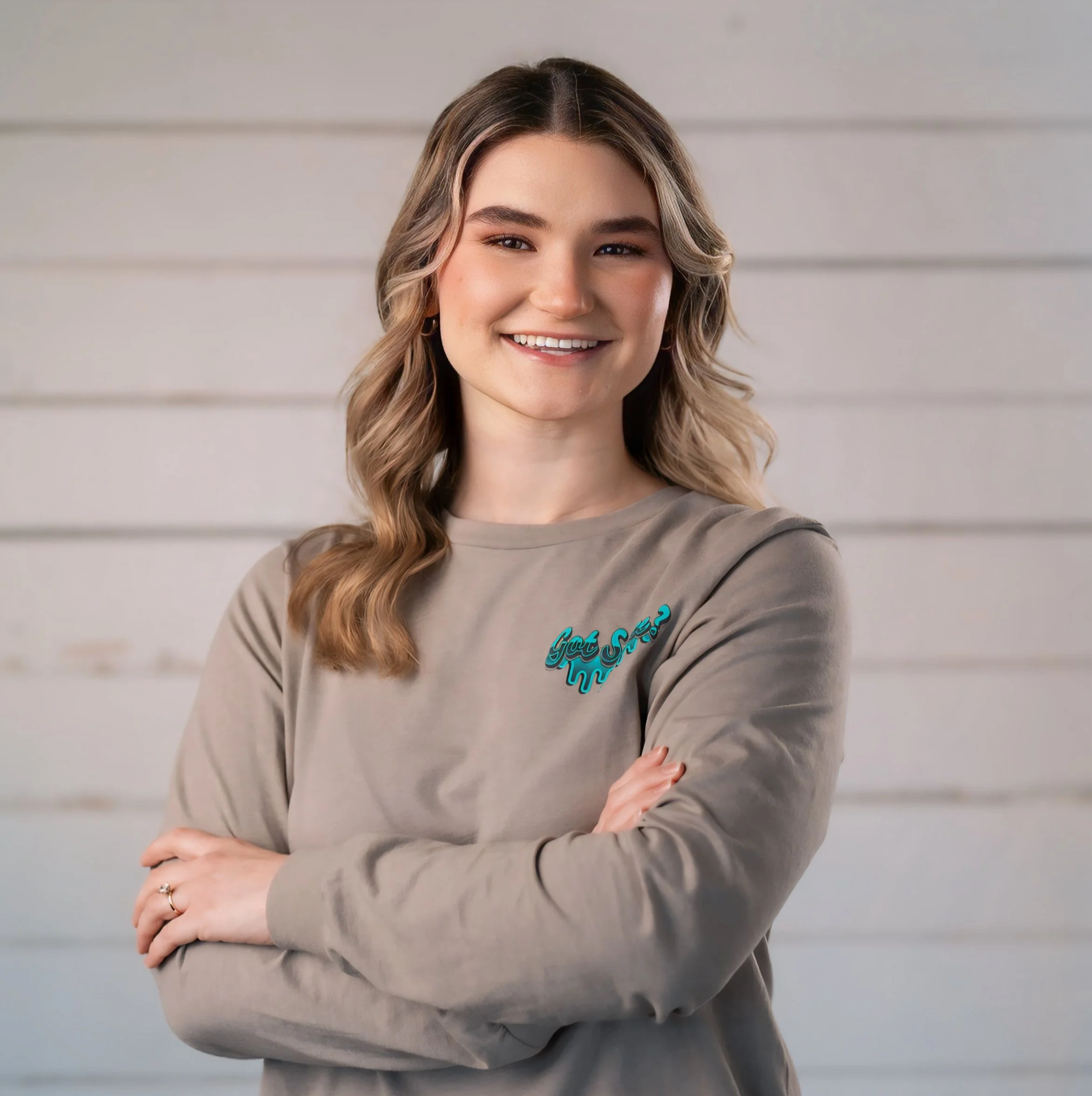 A young woman with wavy blonde hair smiling, standing with arms crossed in front of a white wooden wall, wearing a beige sweatshirt with teal text or design on the chest.