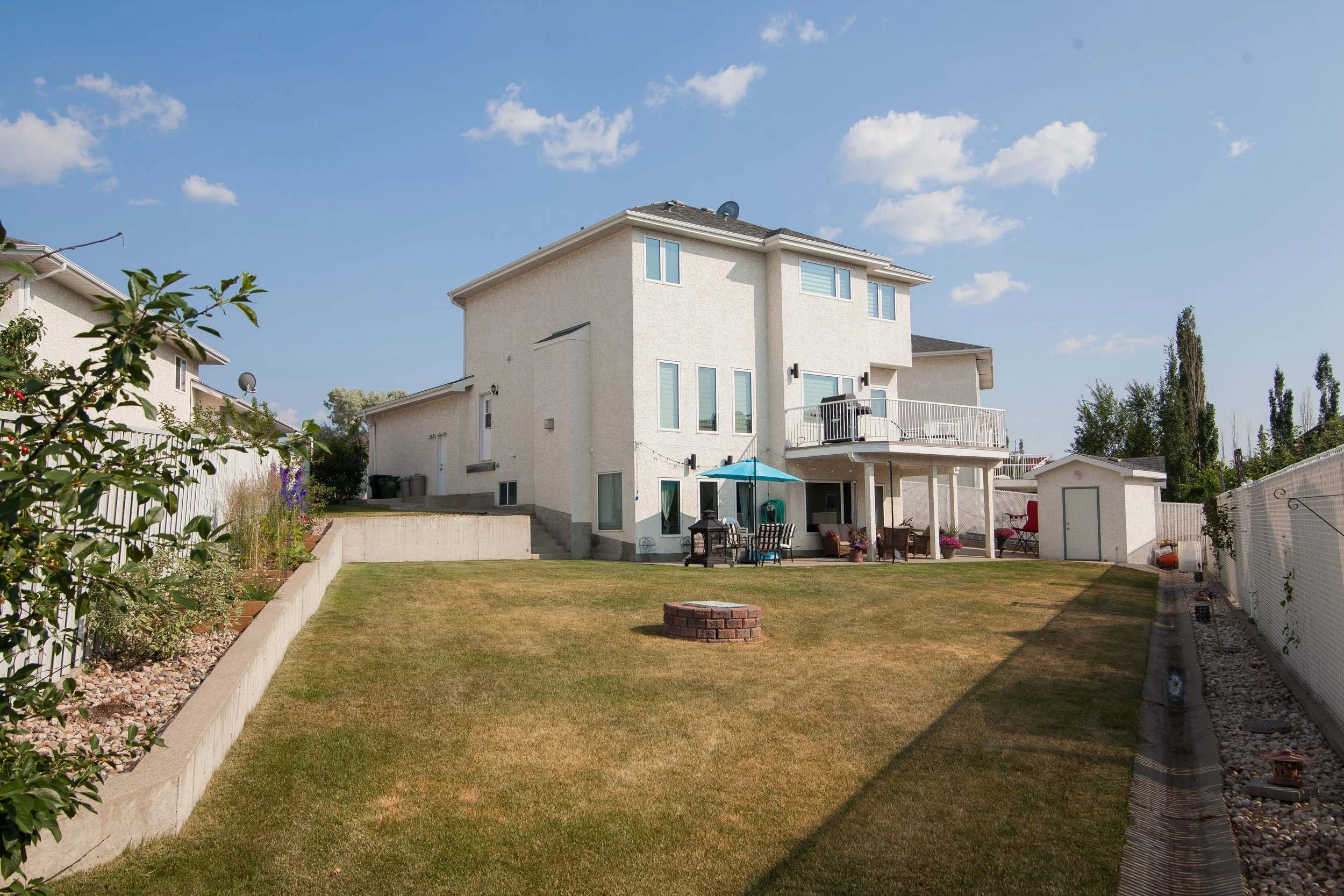 Backyard view of a white three-story house with porch and balcony, grass lawn with a small brick fire pit, outdoor furniture, and a shed, under a blue sky with clouds.