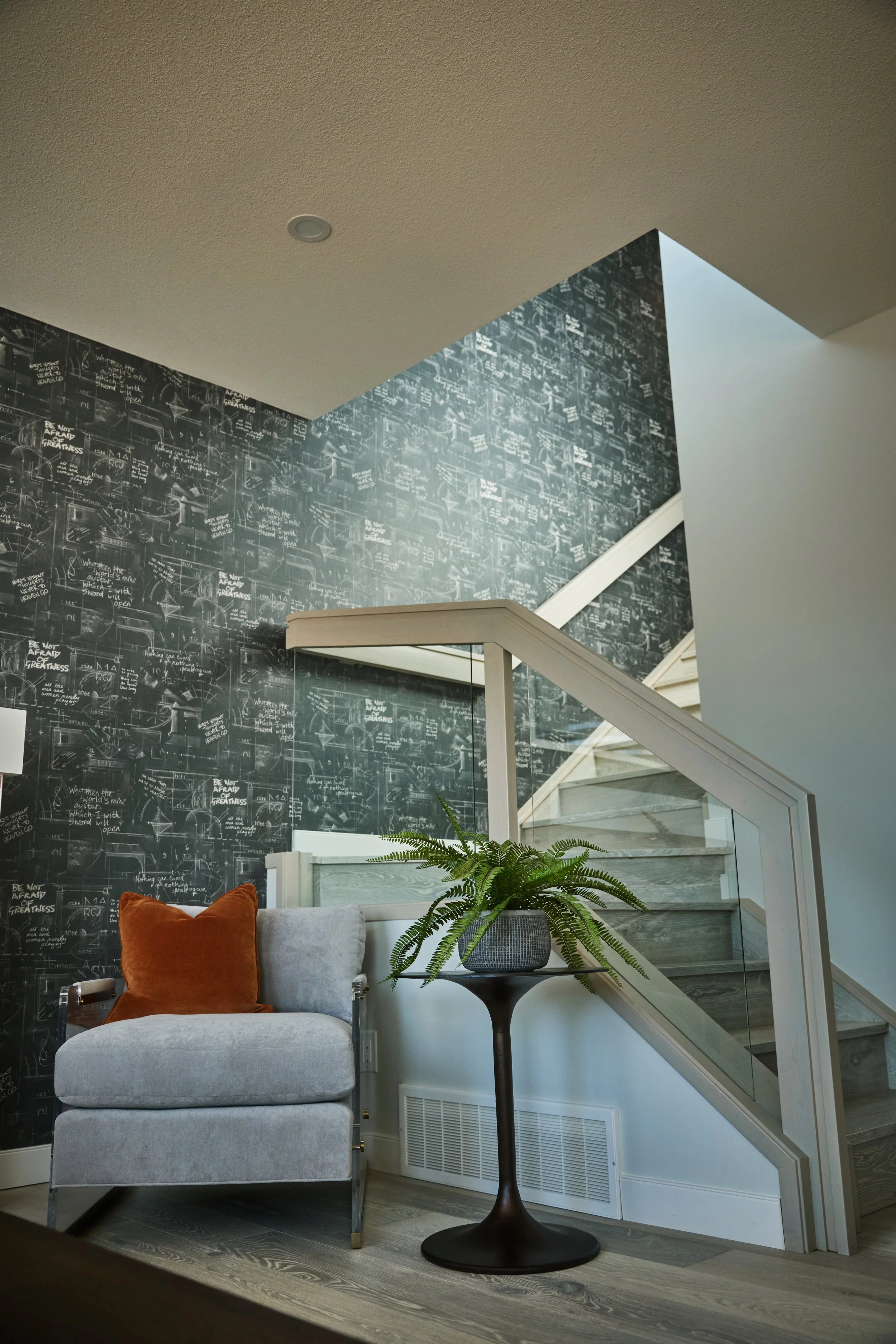 Interior of a home featuring a staircase with glass railing, dark patterned chalkboard wall, gray armchair with orange pillow, a potted fern on a black side table, and wooden flooring.