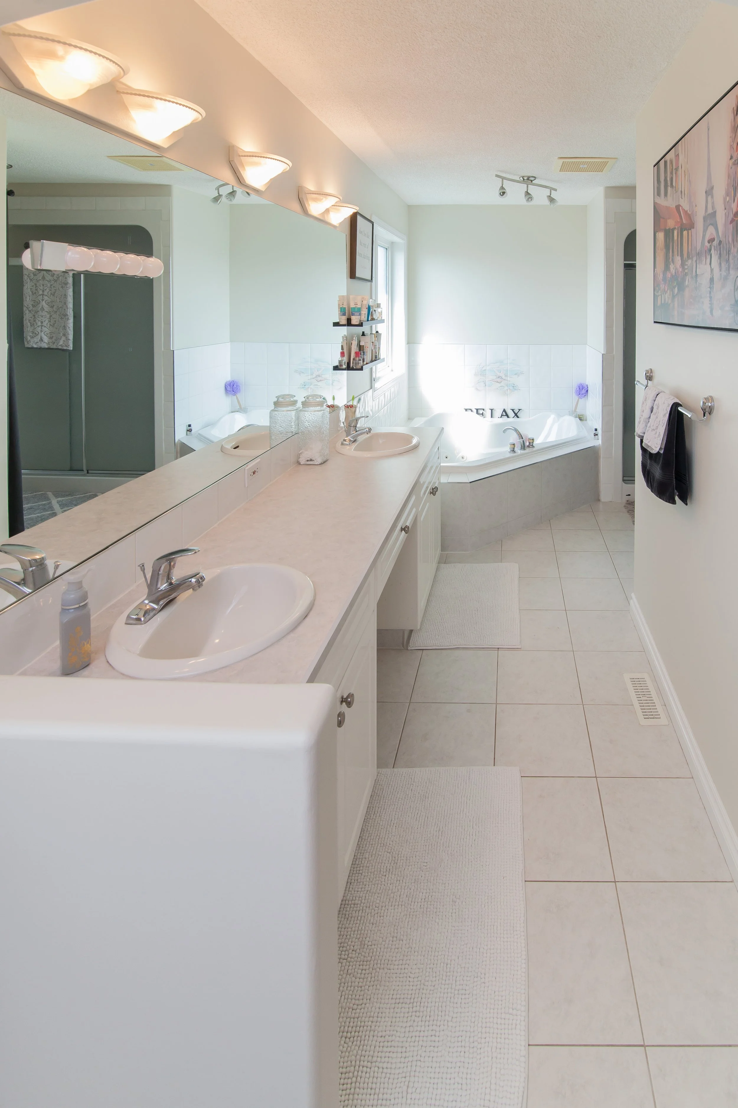 Bathroom with double vanity, large mirror, and a corner bathtub with the word 'RELAX' written on it, decorated with purple flowers, and a walk-in shower in the background.