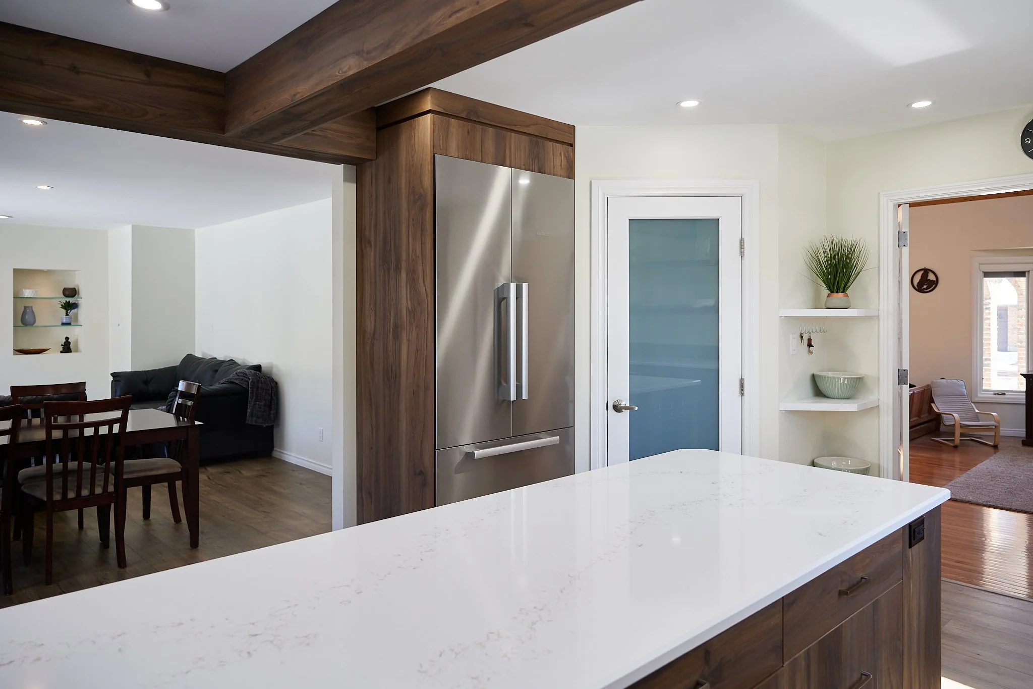 Modern kitchen with white marble countertop and stainless steel refrigerator, view into dining and living area with wood flooring and furniture, potted plant on shelf.