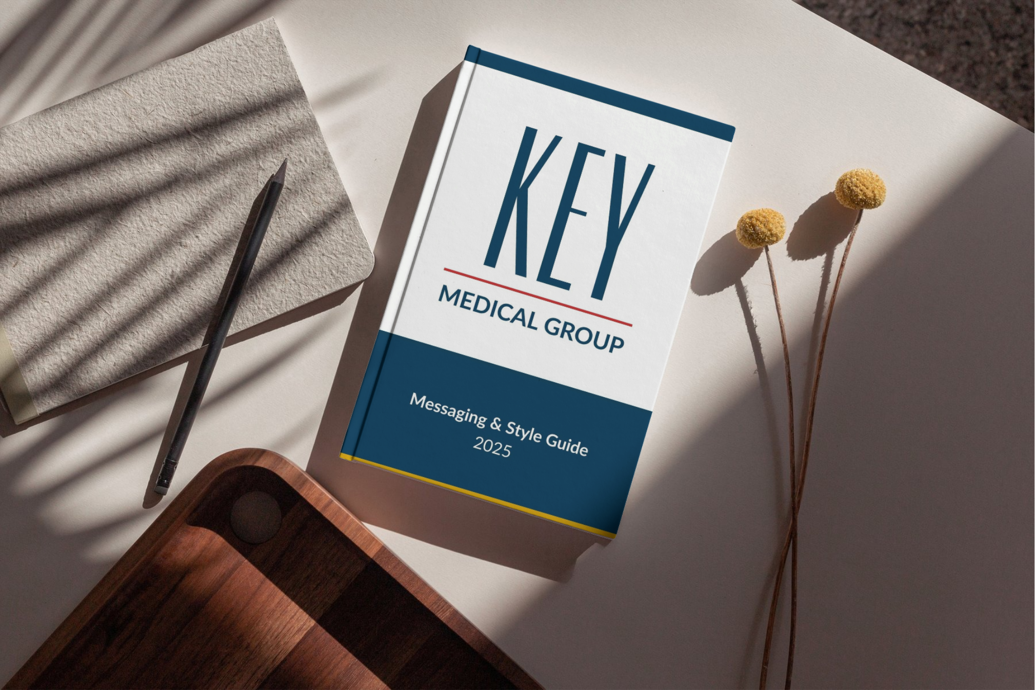 A medical group guidebook, a pen, two yellow spherical flowers, and a textured piece of paper on a white surface with shadows.
