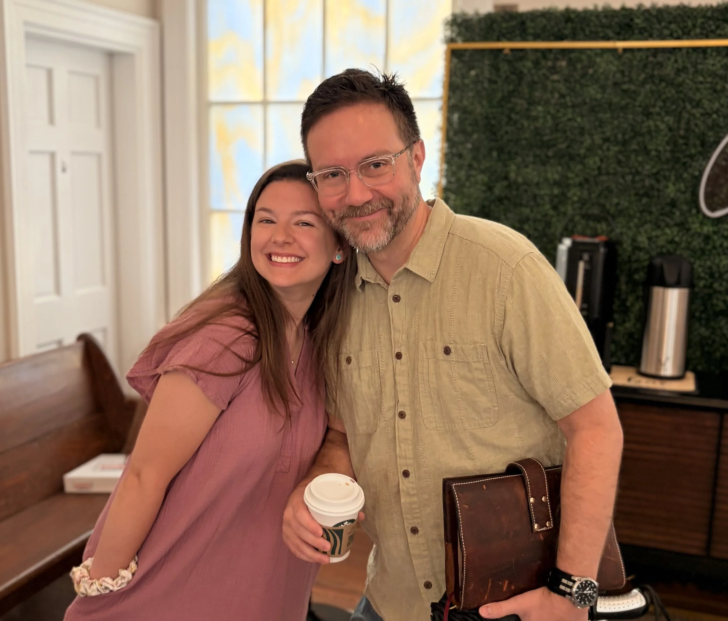 A young woman and an older man are smiling and hugging in a room with a green wall and colorful window art. The woman is holding a coffee and wearing a pink shirt, while the man is holding a brown leather briefcase and a laptop. Both appear happy.