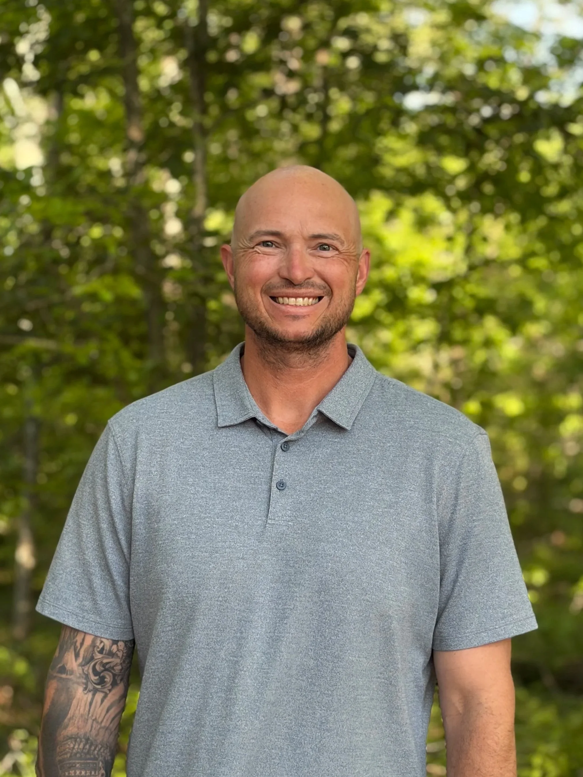 A man standing outdoors, smiling at the camera. The man is bald with a beard, wearing a patterned short-sleeve shirt, and has tattoos on his arms. The woman has long brown hair, tattoos on her arms, and is wearing a sleeveless top and striped pants.