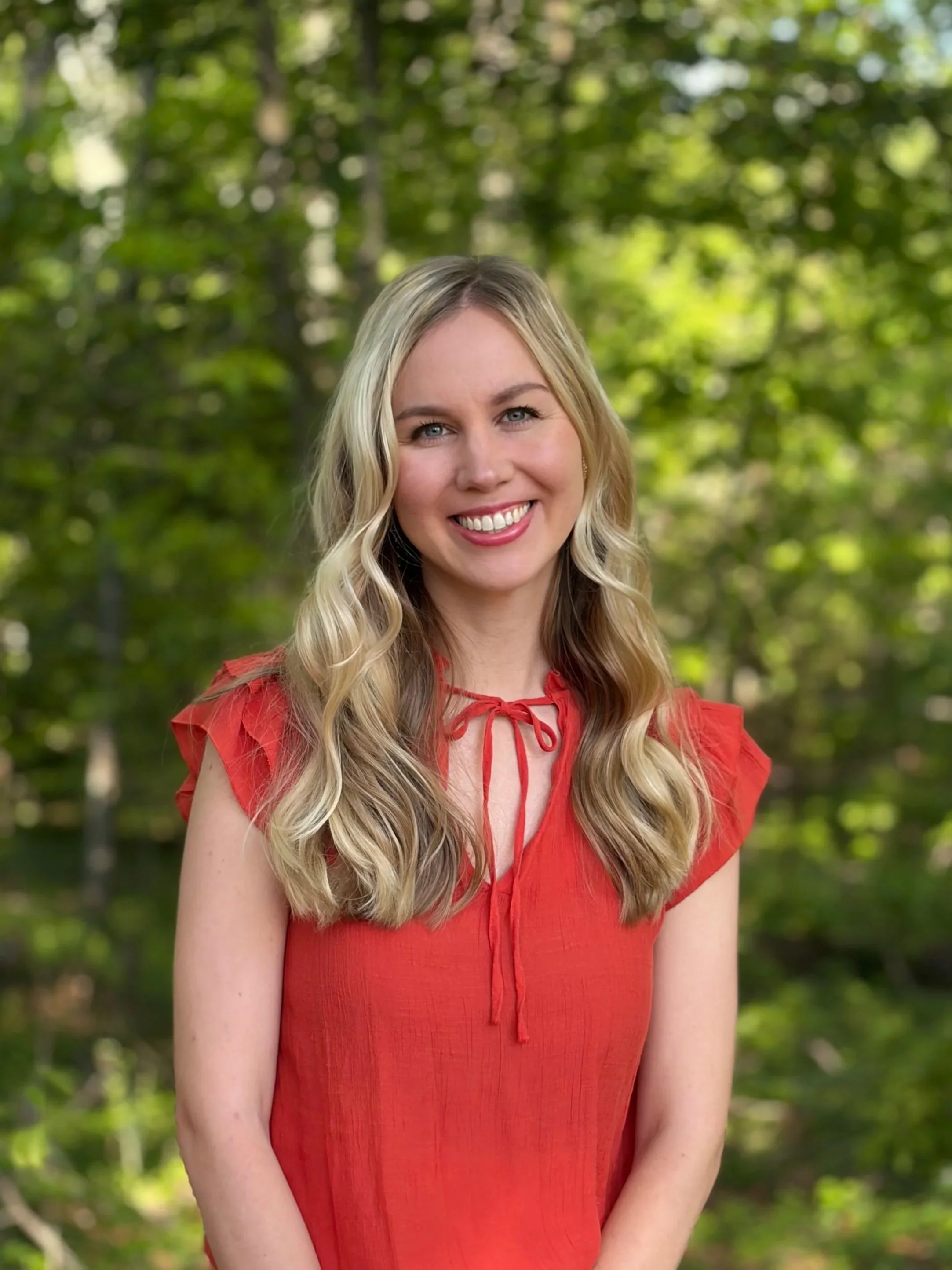 A young woman with long wavy blonde hair, wearing a colorful floral dress with yellow, purple, green, and navy blue patterns, smiling and standing in front of a plain light-colored wall.