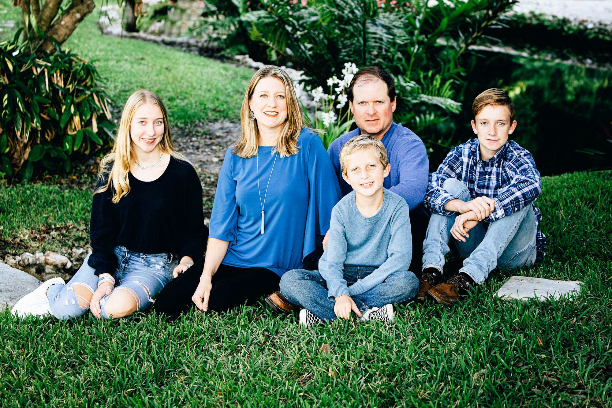 Family posing outdoors with greenery in the background, five people sitting on grass.