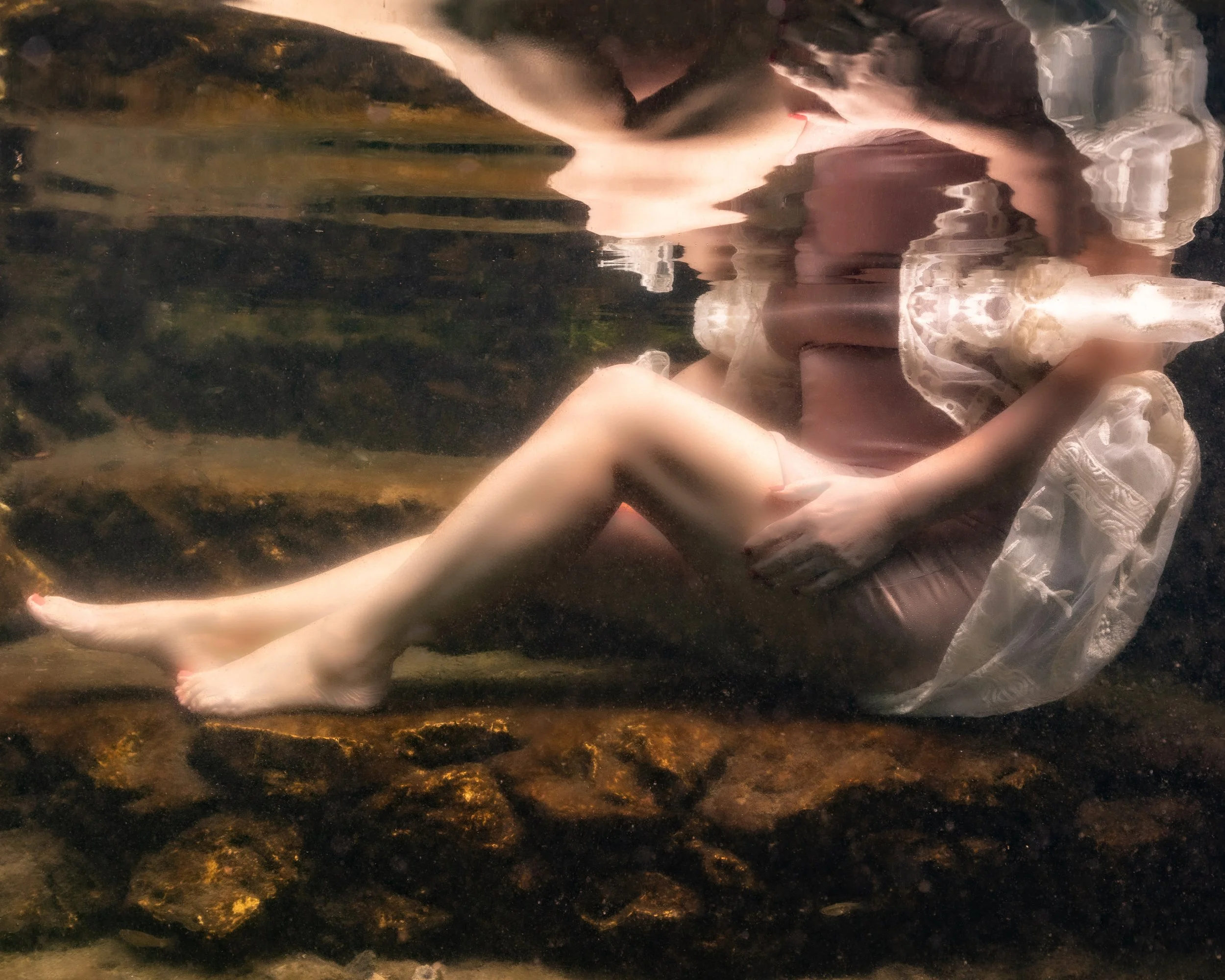 A person submerged underwater, sitting on rocks with legs extended and arms crossed over their knees, wearing a flowing, lace-embellished dress.