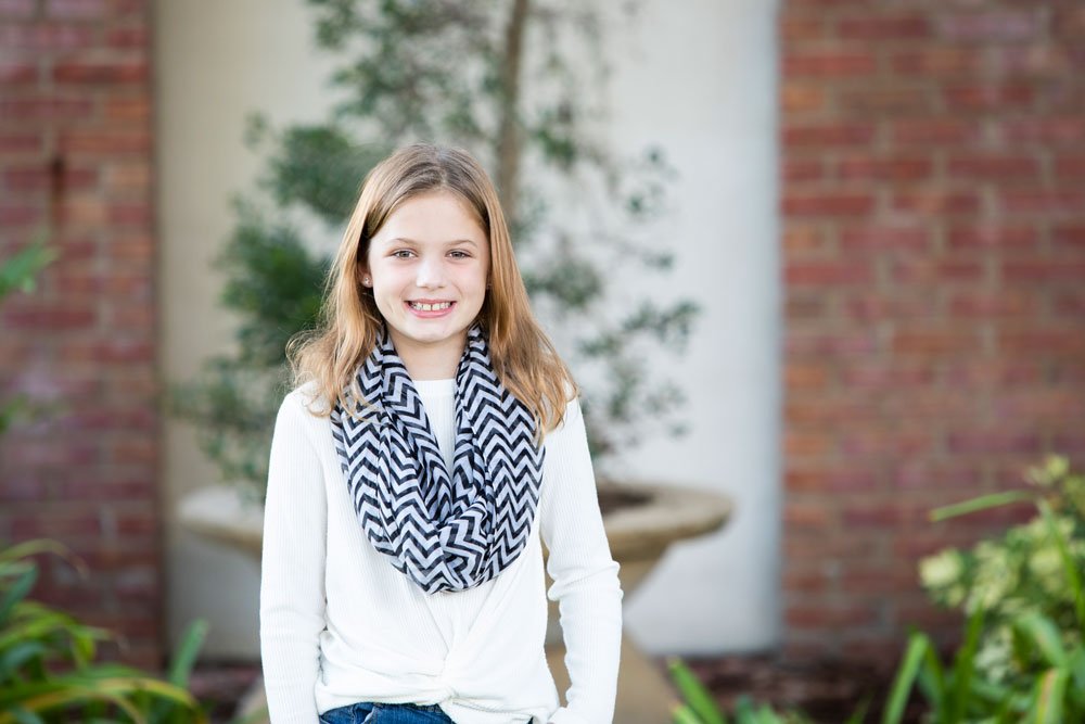 Girl in white sweater with chevron scarf standing outside in garden with brick wall background.