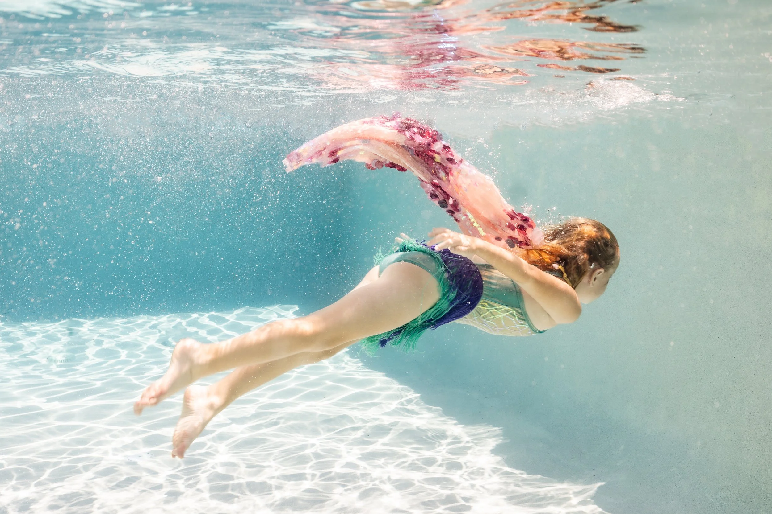 Young girl swimming underwater in a pool, wearing a colorful swimsuit, with a pink and purple fish-shaped swim fin.