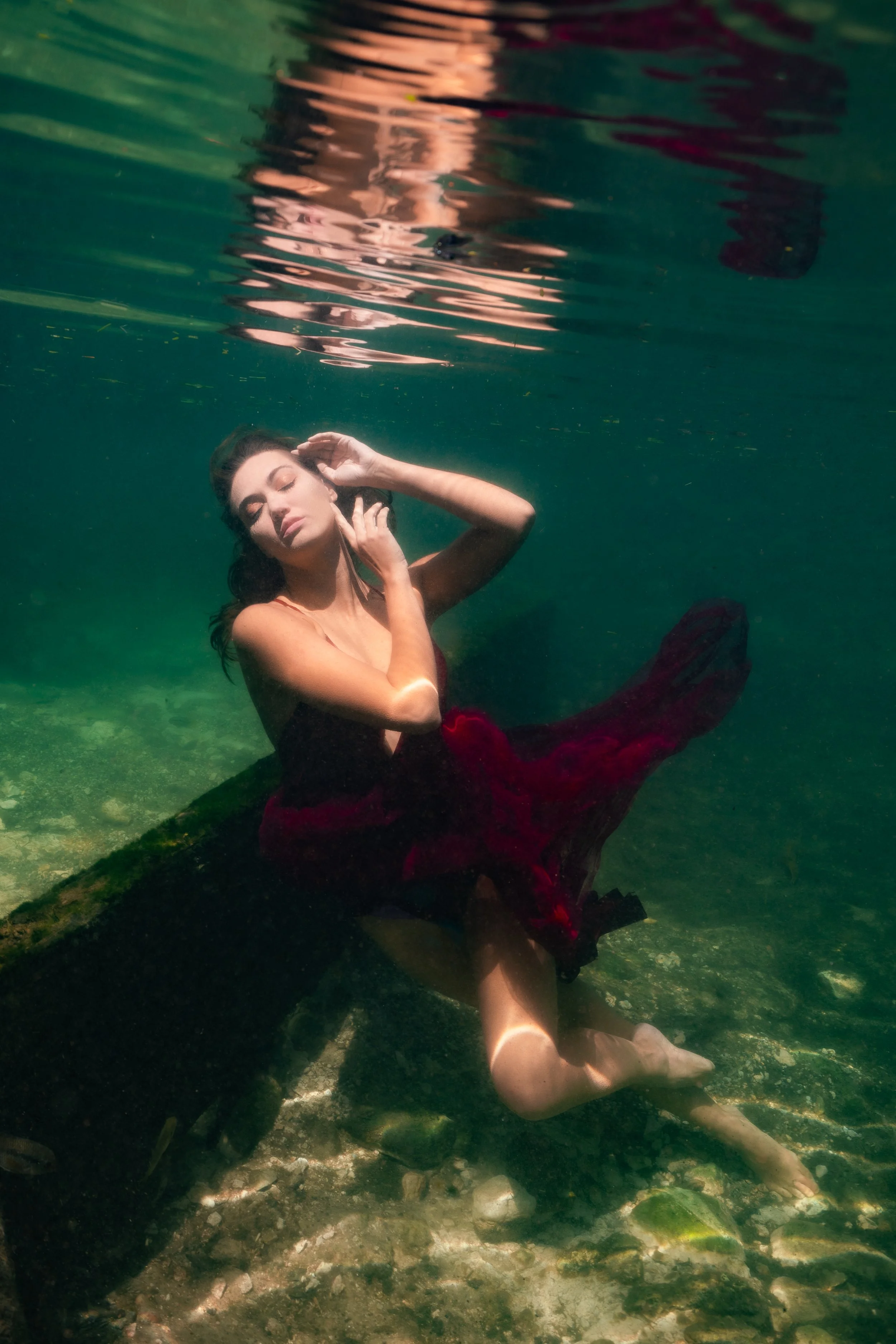 A woman sitting underwater on a submerged log, wearing a dark red dress and posing with her eyes closed and one hand near her face.
