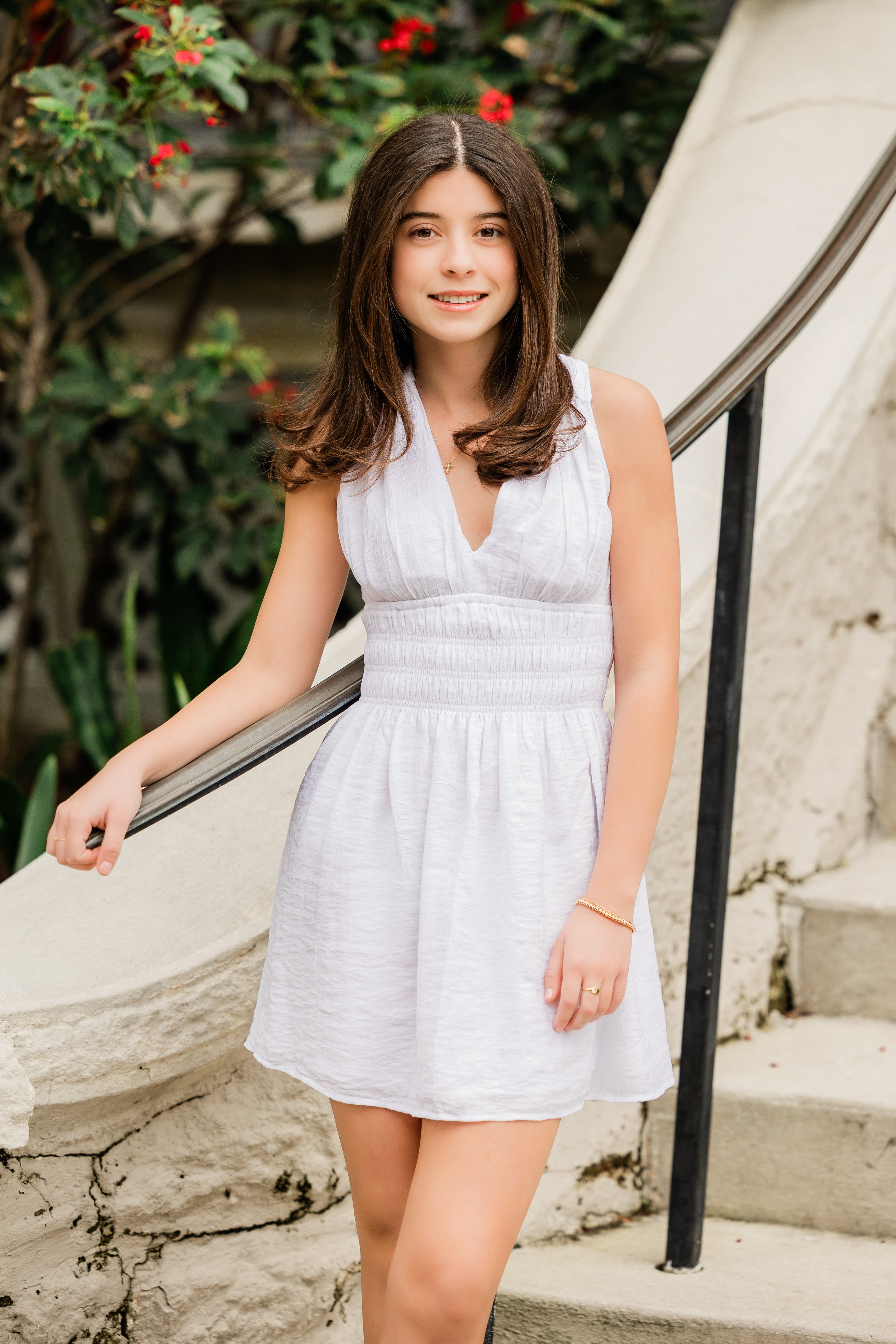 Young woman in white dress posing by stairs and bushes with red flowers