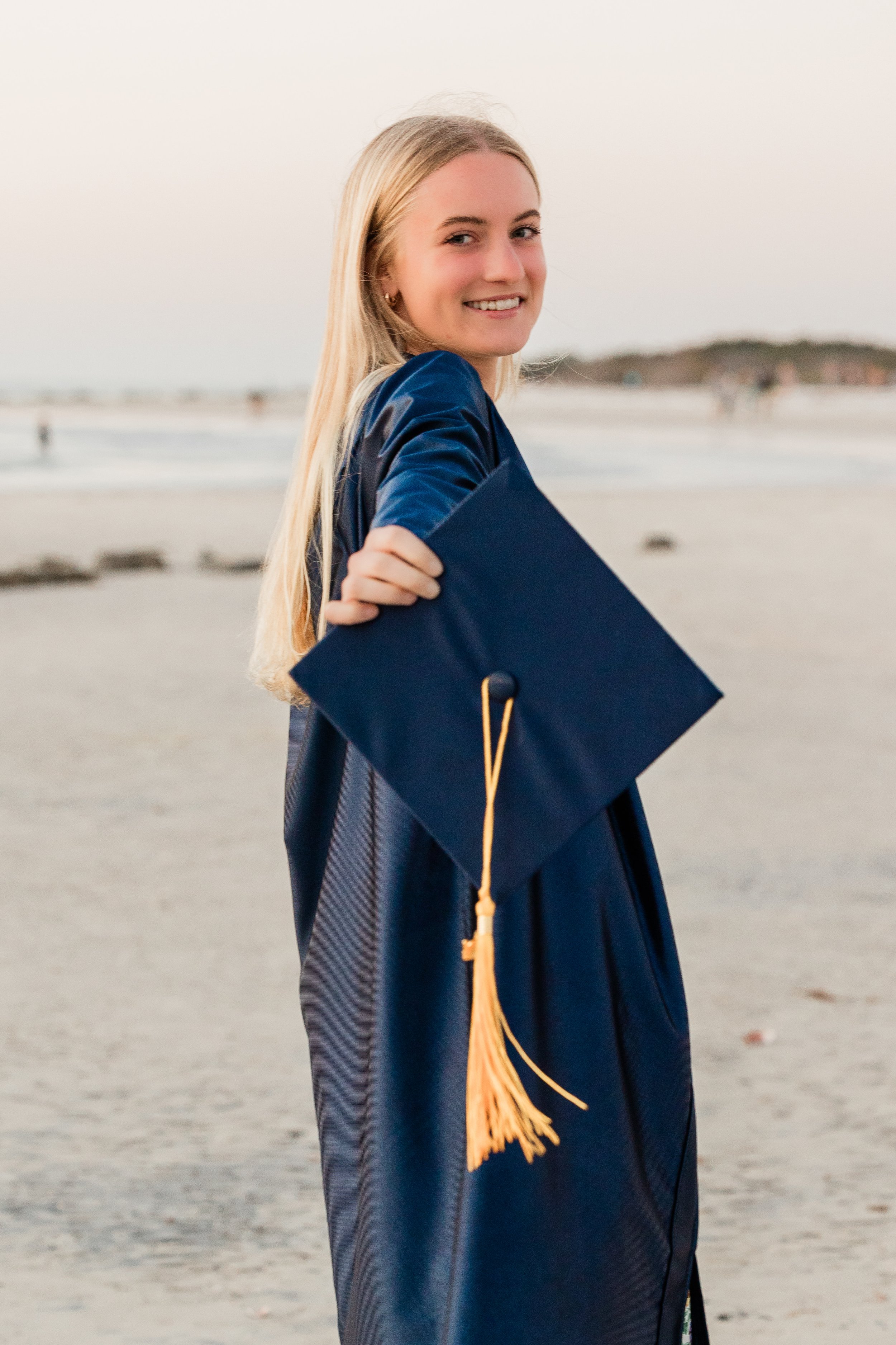 Young woman in graduation gown holding a cap at the beach.