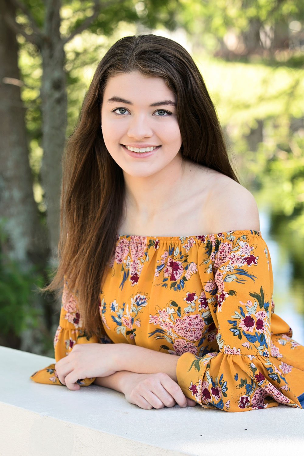 Young woman with long brown hair wearing a floral off-the-shoulder top, leaning on a white surface, smiling, with a blurred green background.
