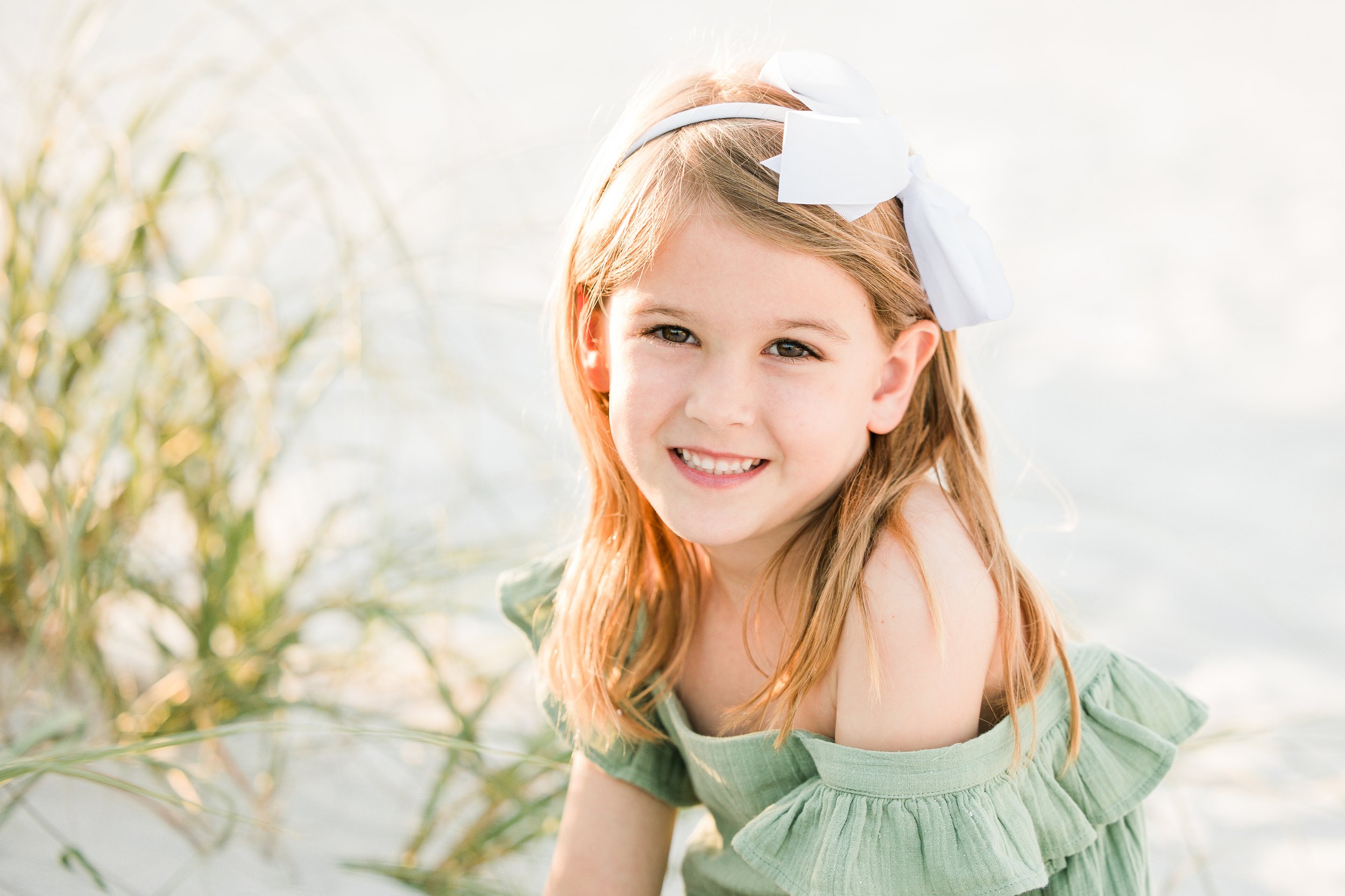 Young girl with a white bow and green dress, smiling outdoors.