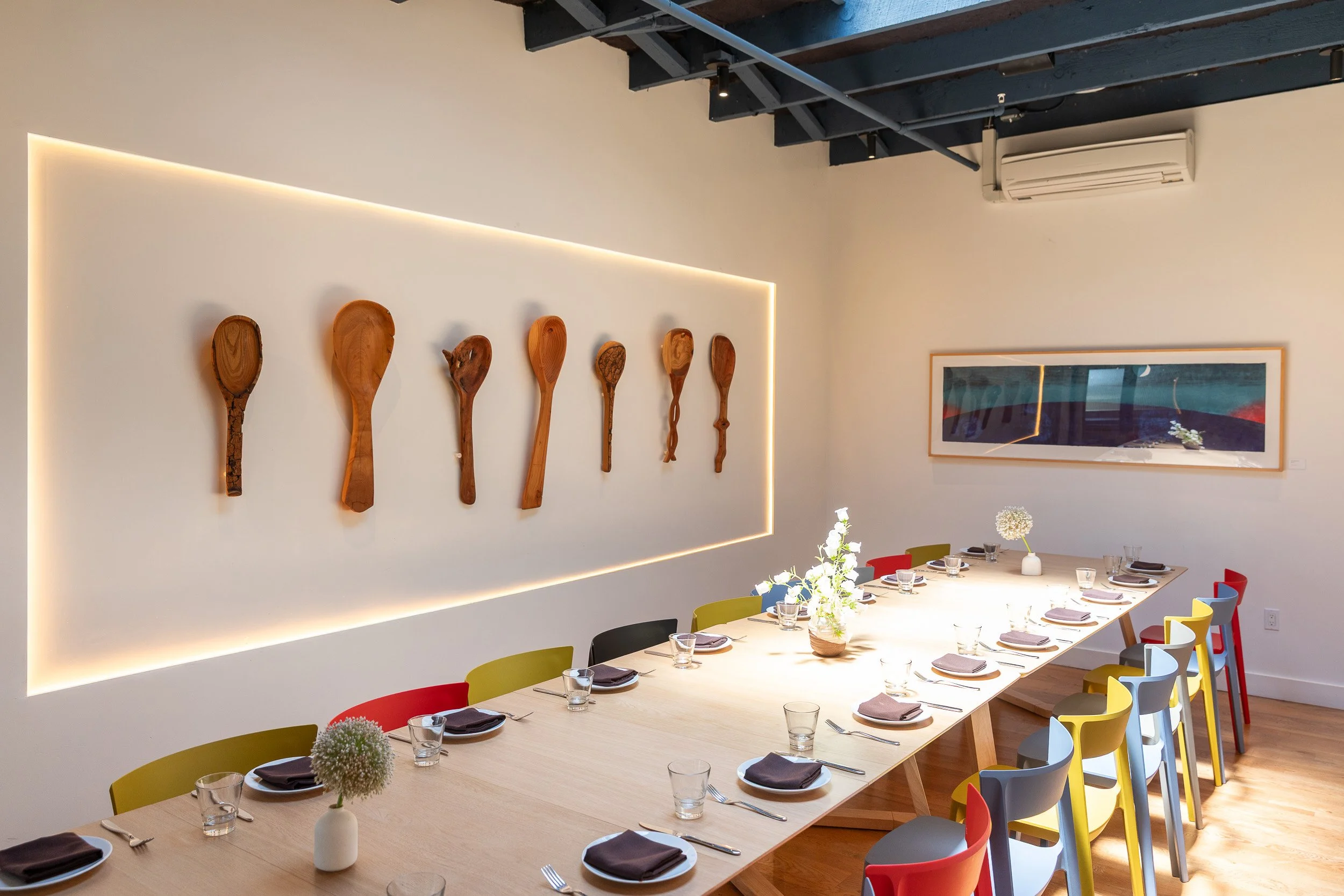 A modern dining room with a long gray table, colorful chairs, and decorative wooden spoons on the wall.