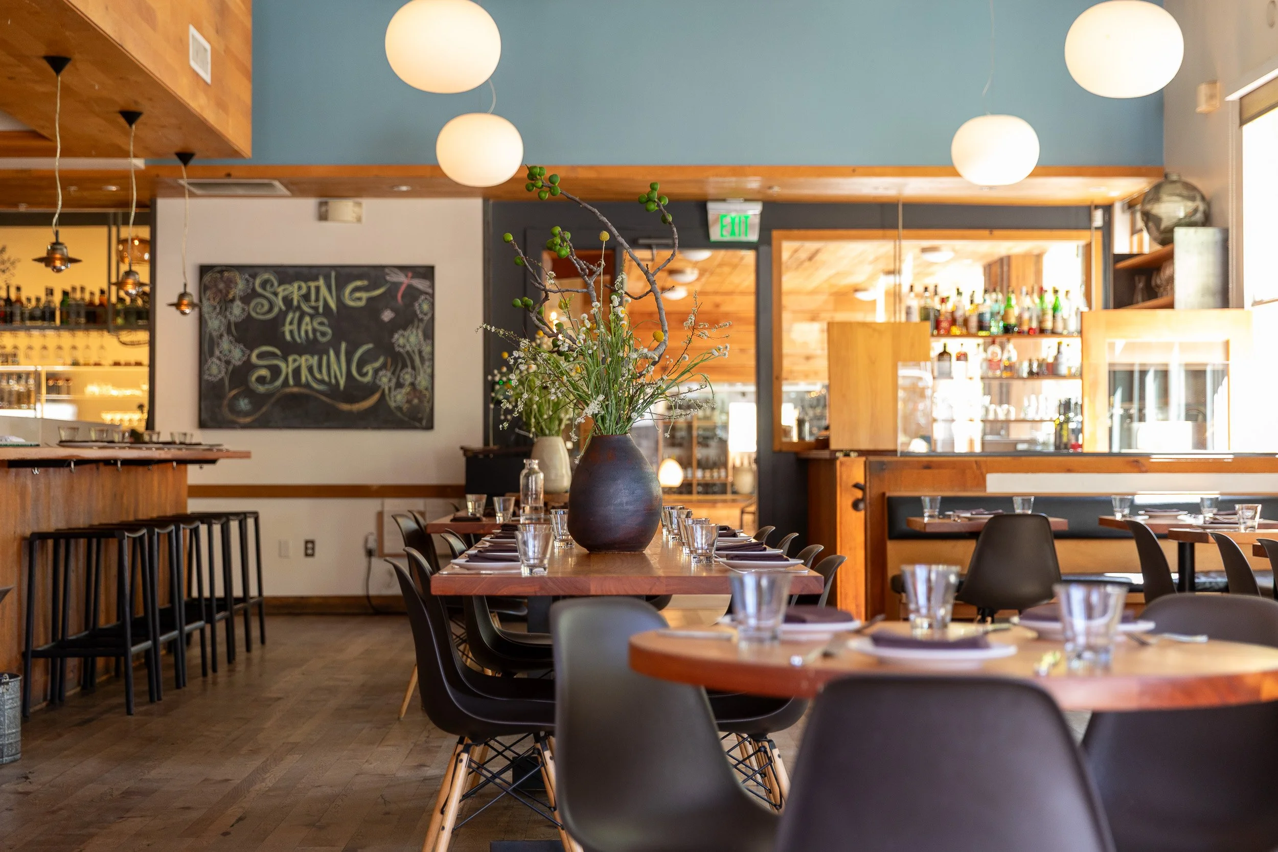 Interior of a restaurant with a wooden table set with glasses and napkins, a large vase with branches and green berries as a centerpiece, hanging globe lights, a chalkboard wall, and bar shelves in the background.